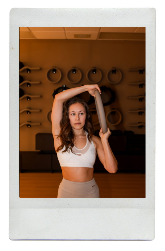 A young woman with long brown hair doing a fitness pose with a foam roller in a gym.