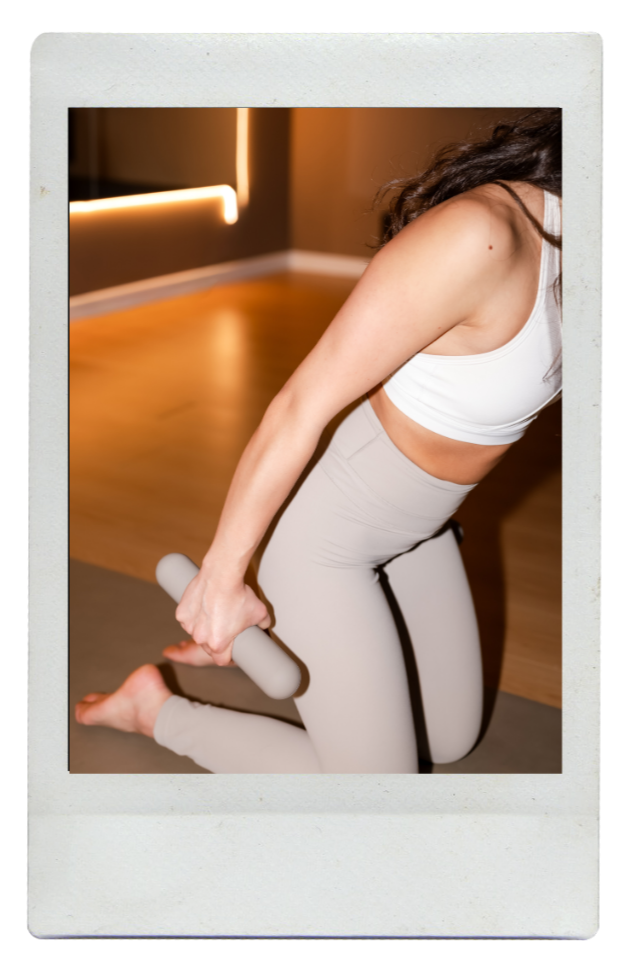 A woman in athletic clothing kneeling on a yoga mat, holding a massage roller in her right hand, in a room with hardwood floors and warm lighting.