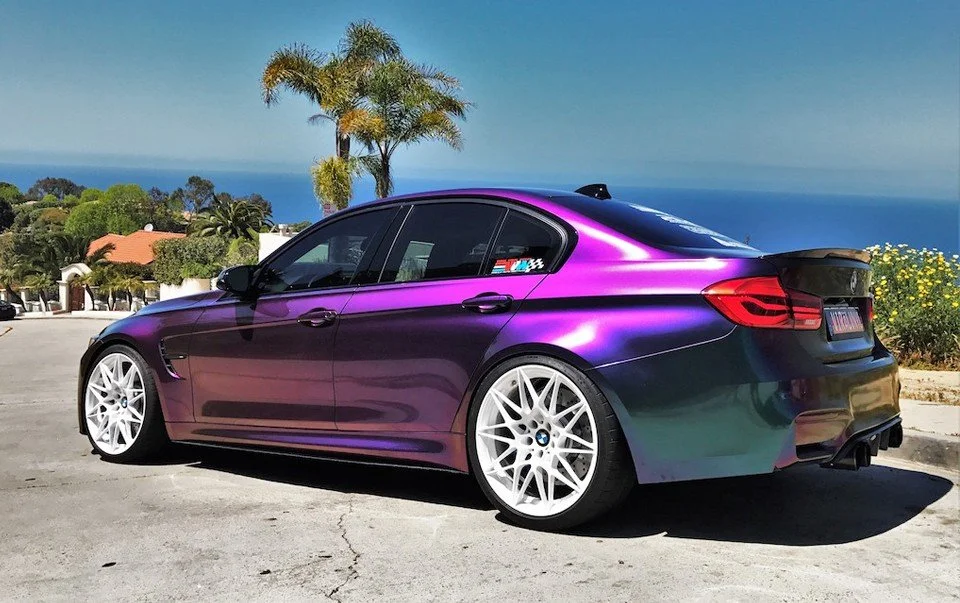 A purple and black BMW sedan with custom white wheels parked outdoors near palm trees and ocean view.
