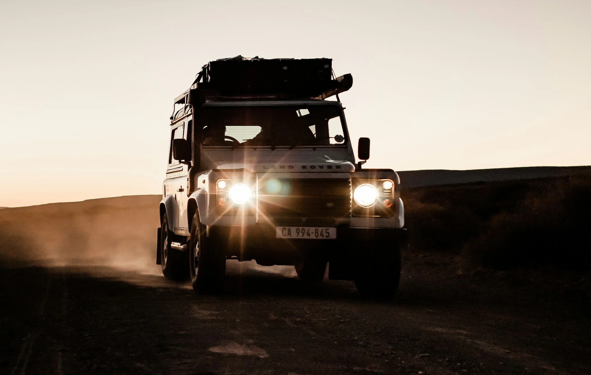 A classic Land Rover driving on a dirt road at sunset, kicking up dust.