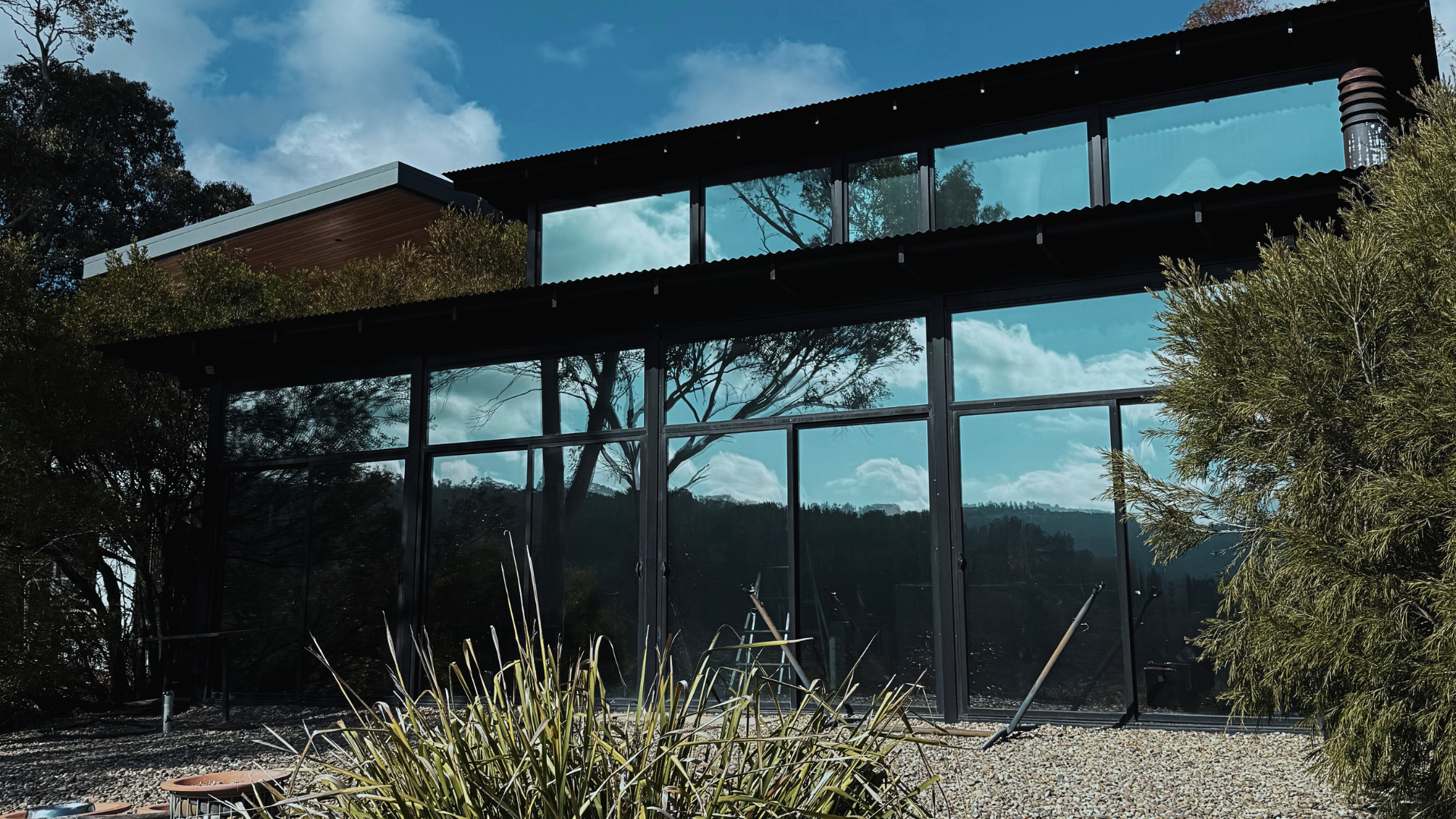 Modern house with large glass windows, black framing, and a sloped roof. Surrounded by trees and plants under a partly cloudy sky.