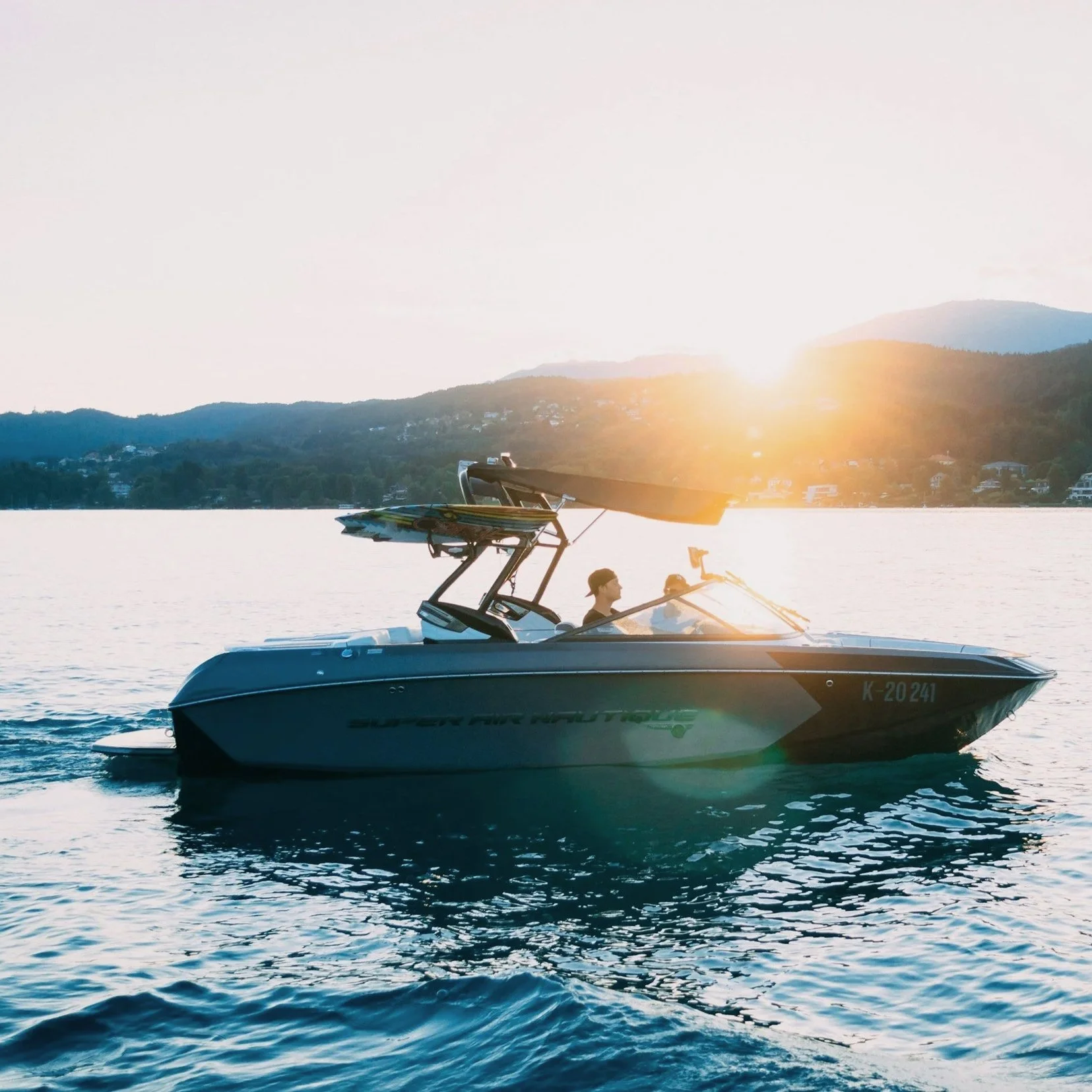 A speedboat on a lake with two people on it, sailing during sunset with a mountain and houses in the background.
