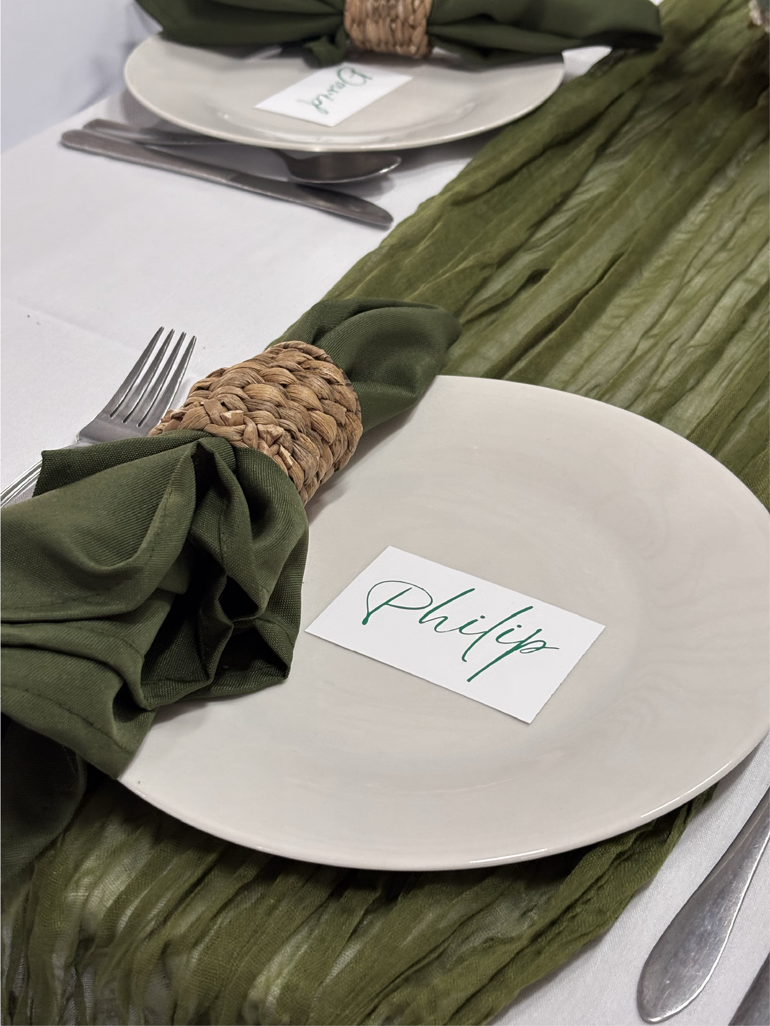 A place setting at a formal dinner with white plates, silverware, and a green cloth napkin tied with a woven napkin ring. A name card with the name 'Philip' is placed on the plate, and there is a green table runner underneath.