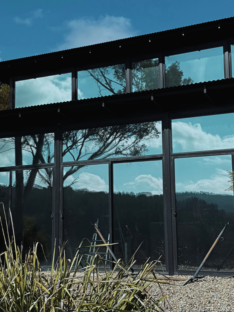 A modern building with large glass windows reflecting trees and a partly cloudy sky, surrounded by outdoor plants and gravel.