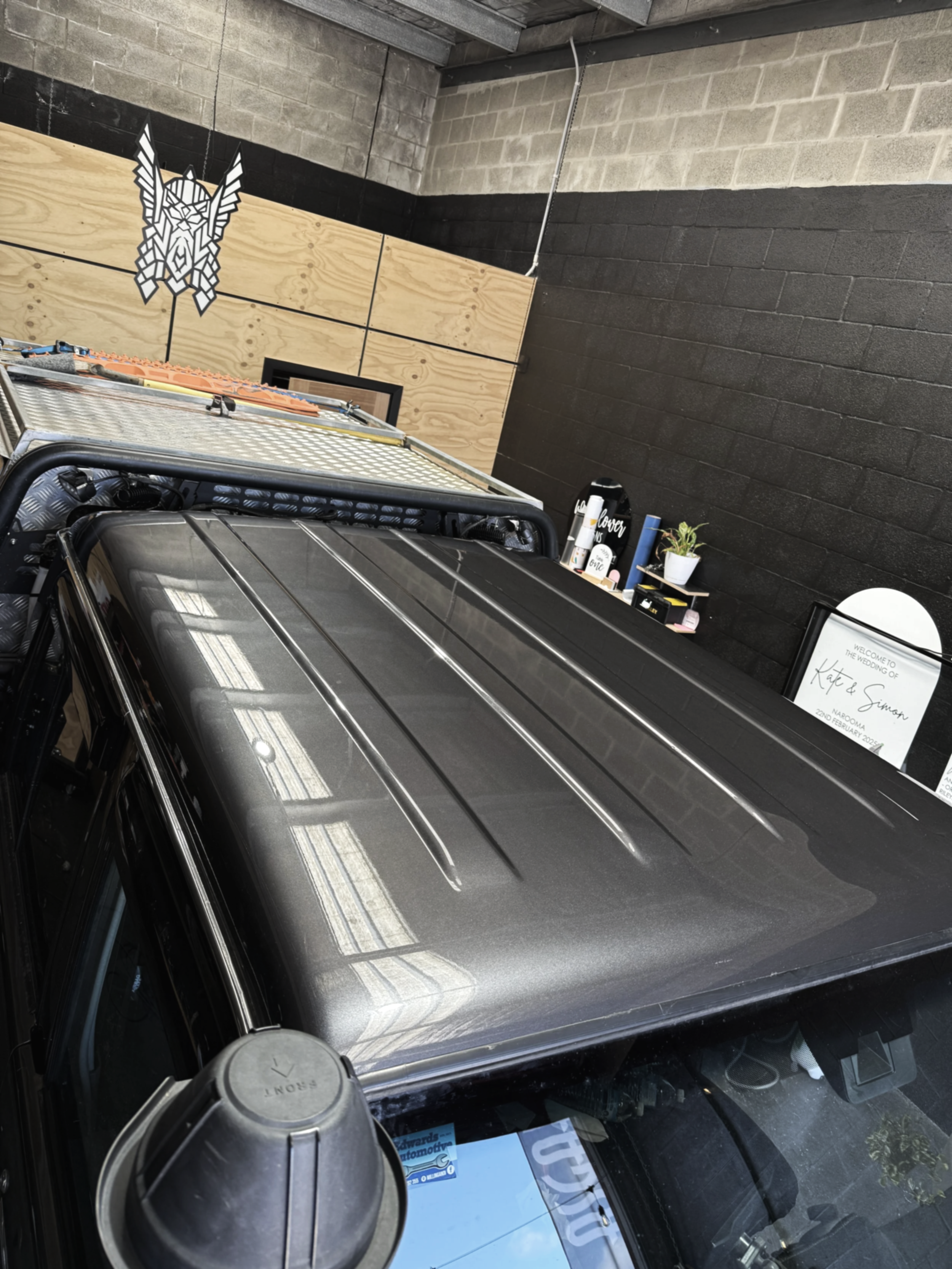 Close-up of a black vehicle with a shiny roof parked indoors, with a partially visible sign in the background that reads 'Welcome to the wedding of Kate & Simon' and a decorated interior wall.