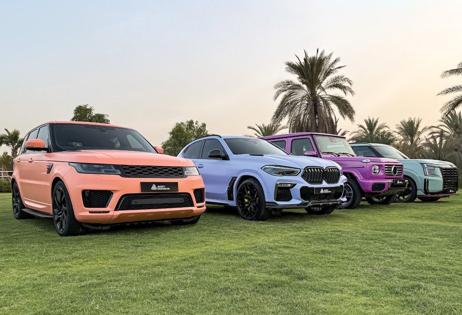 Lineup of five luxury and sports cars on a grassy field with palm trees in the background, featuring a peach-colored Range Rover Sport, a blue BMW X6 M, a purple Mercedes-Benz G-Class, and two other dark-colored SUVs.