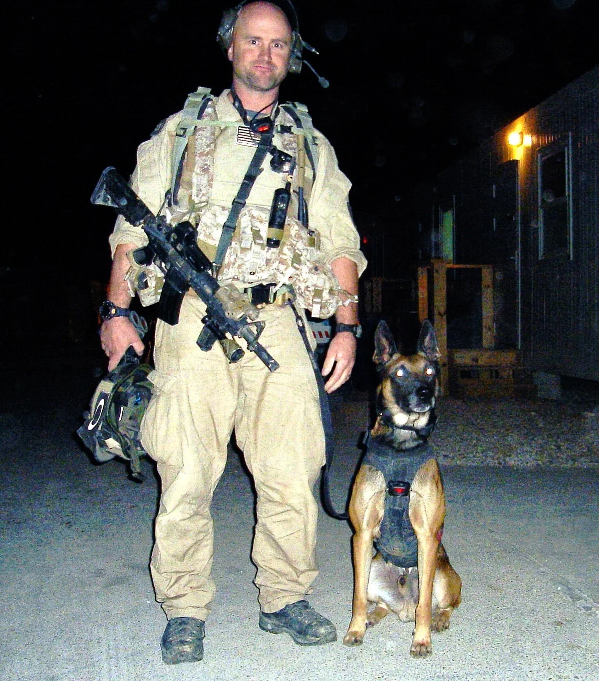 A man in military tactical gear standing outdoors at night with a German Shepherd service dog.