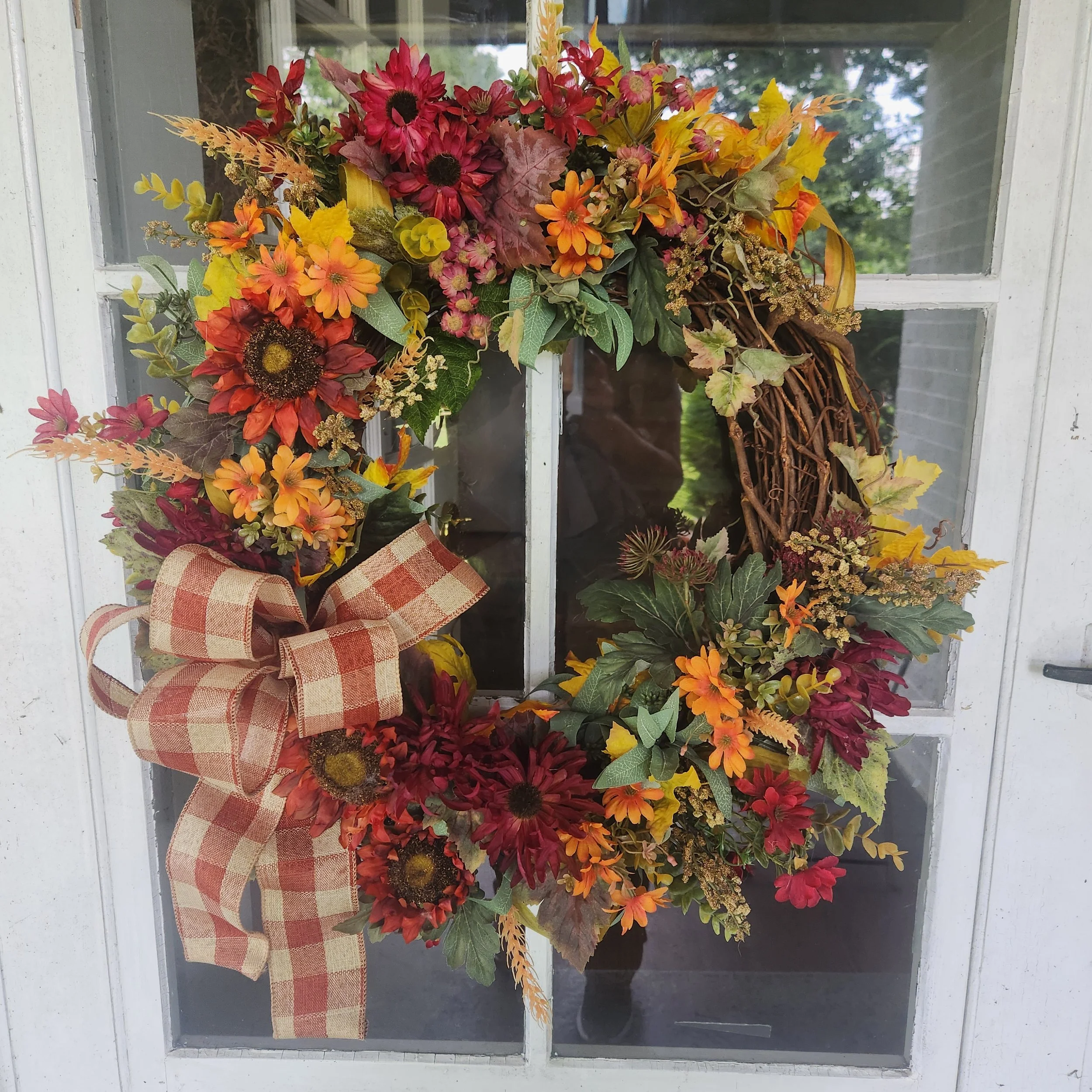 An abundant fall statement piece layered with high-quality silk chrysanthemums, warm autumn leaves, and a classic plaid ribbon. Specially treated to look stunningly fresh all season long.