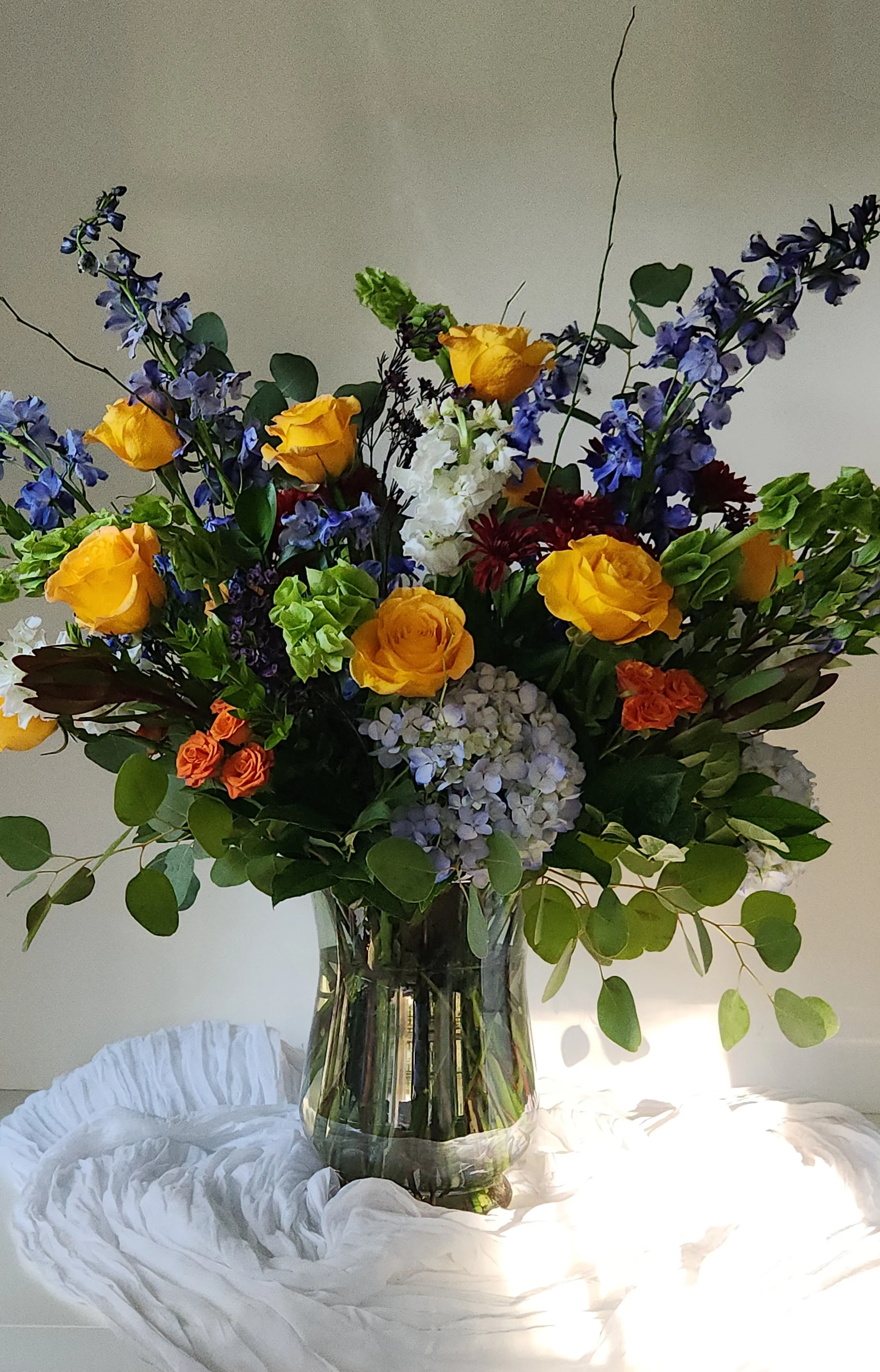 A vibrant and bountifully textured tribute featuring yellow Brighton roses and towering dark blue delphinium. This expressive design was artfully composed as a celebration of life, blending bells of Ireland, fragrant stock, and blue hydrangeas with a