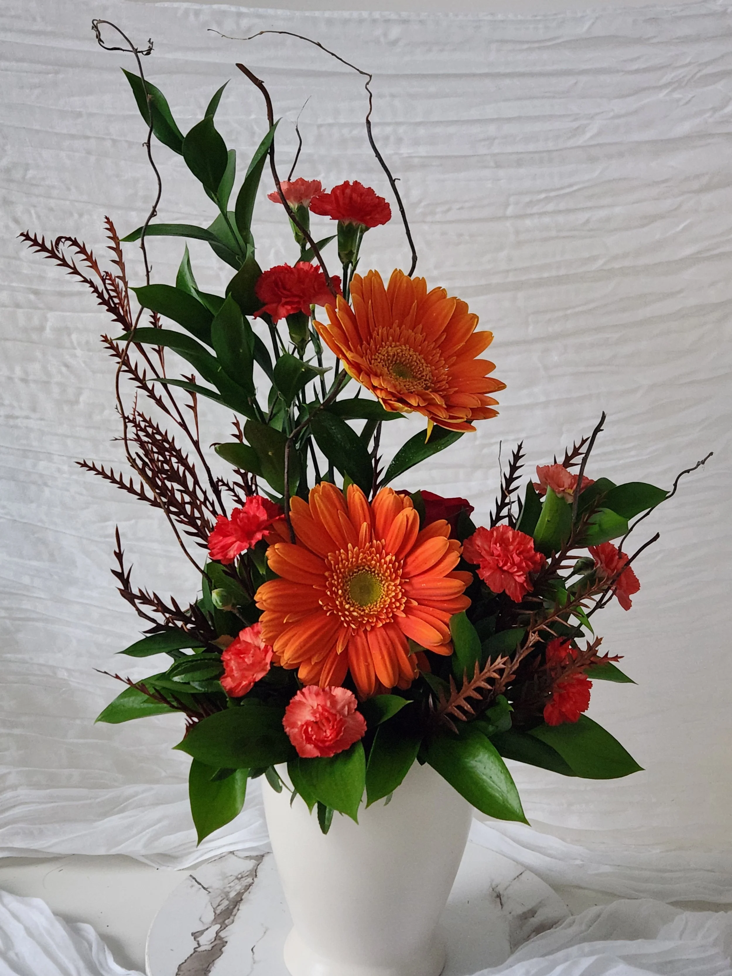 A vibrant, vertical design featuring bold orange gerbera daisies and soft coral mini carnations. Tall, whimsical curly willow branches and deep green foliage create a sense of height and natural movement in a simple white vase