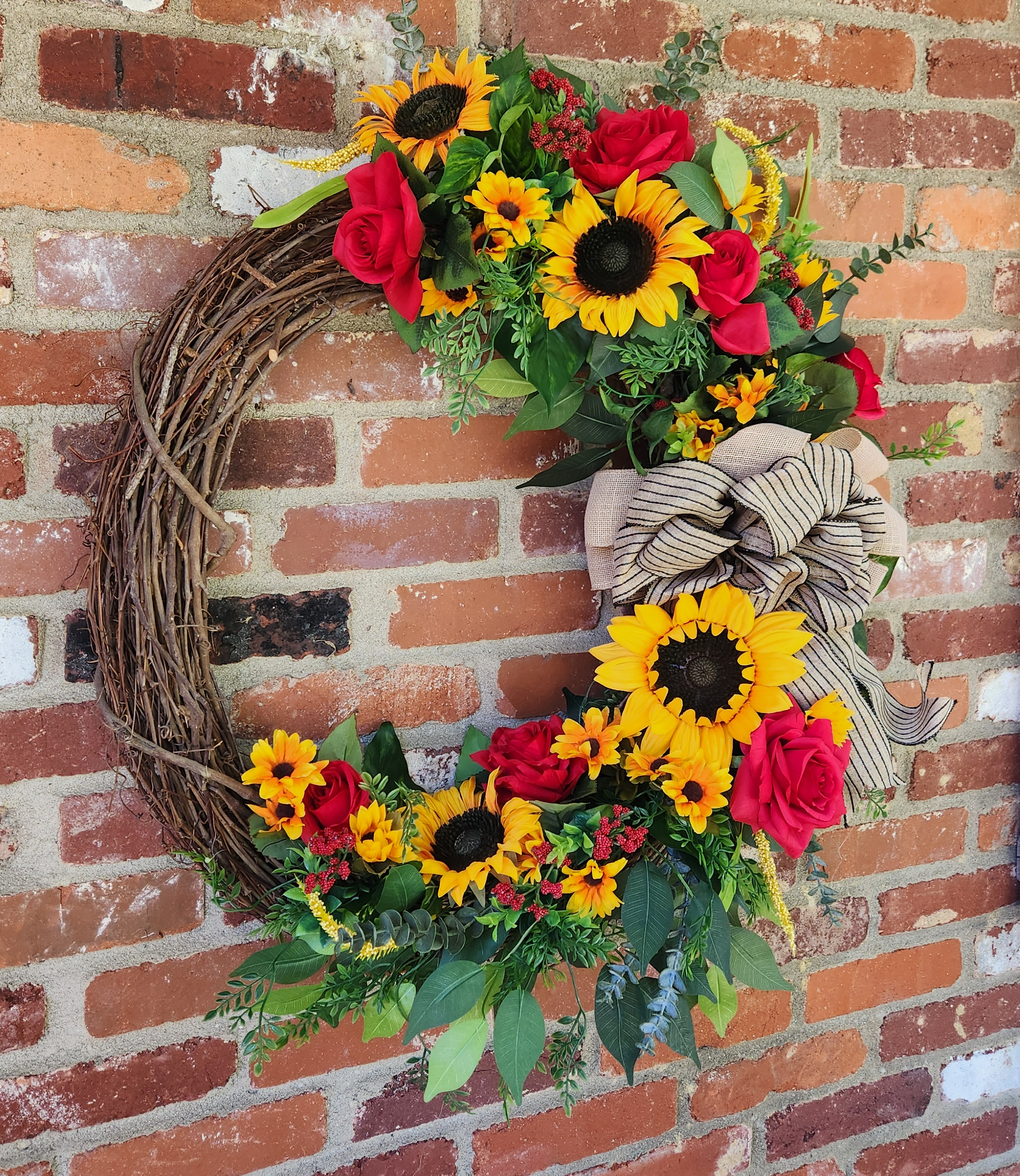 An asymmetrical grapevine wreath featuring vibrant silk sunflowers, rich red roses, and a rustic burlap bow. It brings a gorgeous, welcoming burst of autumn color to any front door or brick entryway.