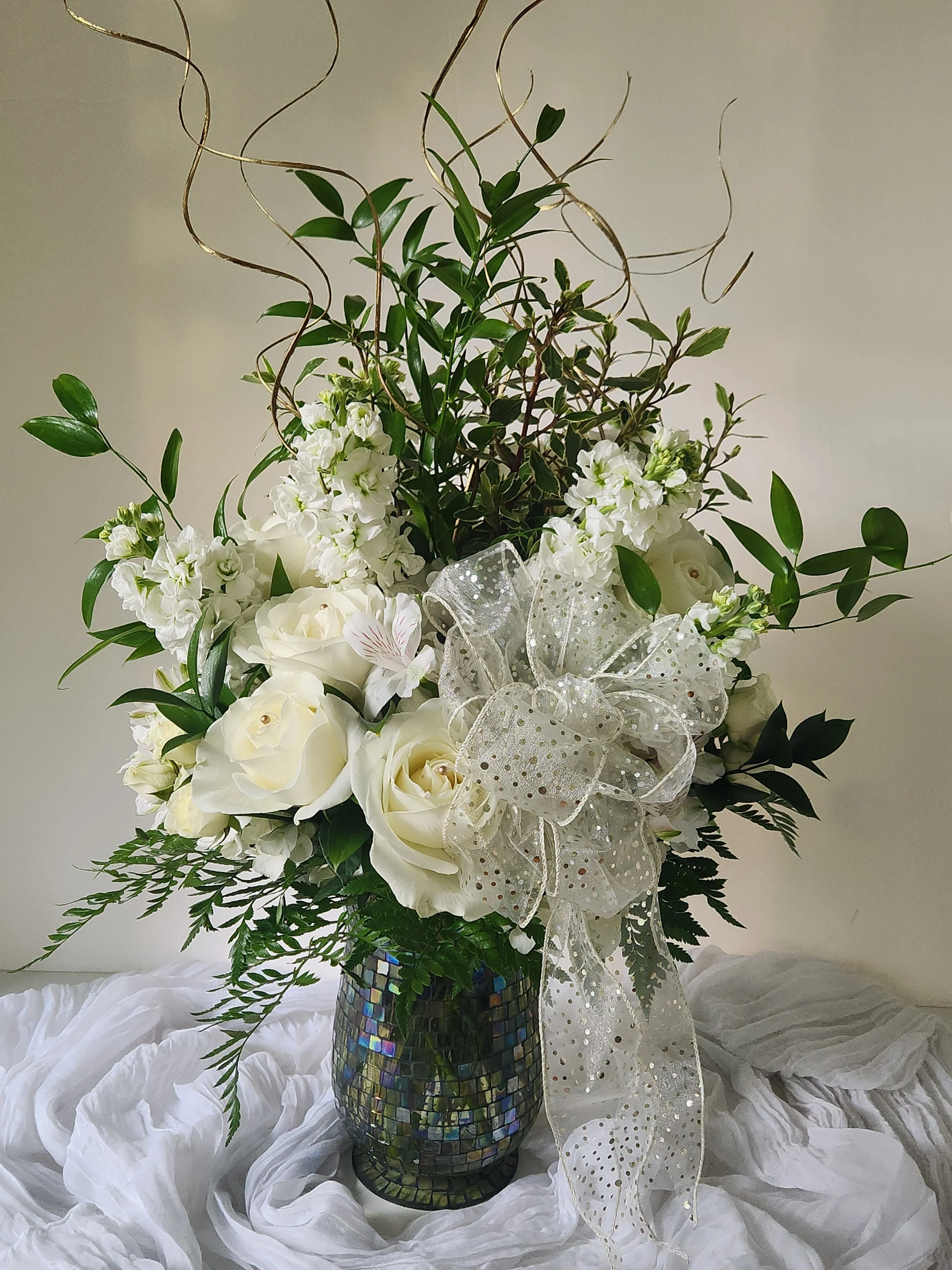 An elegant, all-white design featuring premium roses and the incredible fragrance of fresh white stock. This richly layered arrangement is detailed with gold pearl-tipped accents, shimmering golden branches, and a sheer bow—a sophisticated choice for
