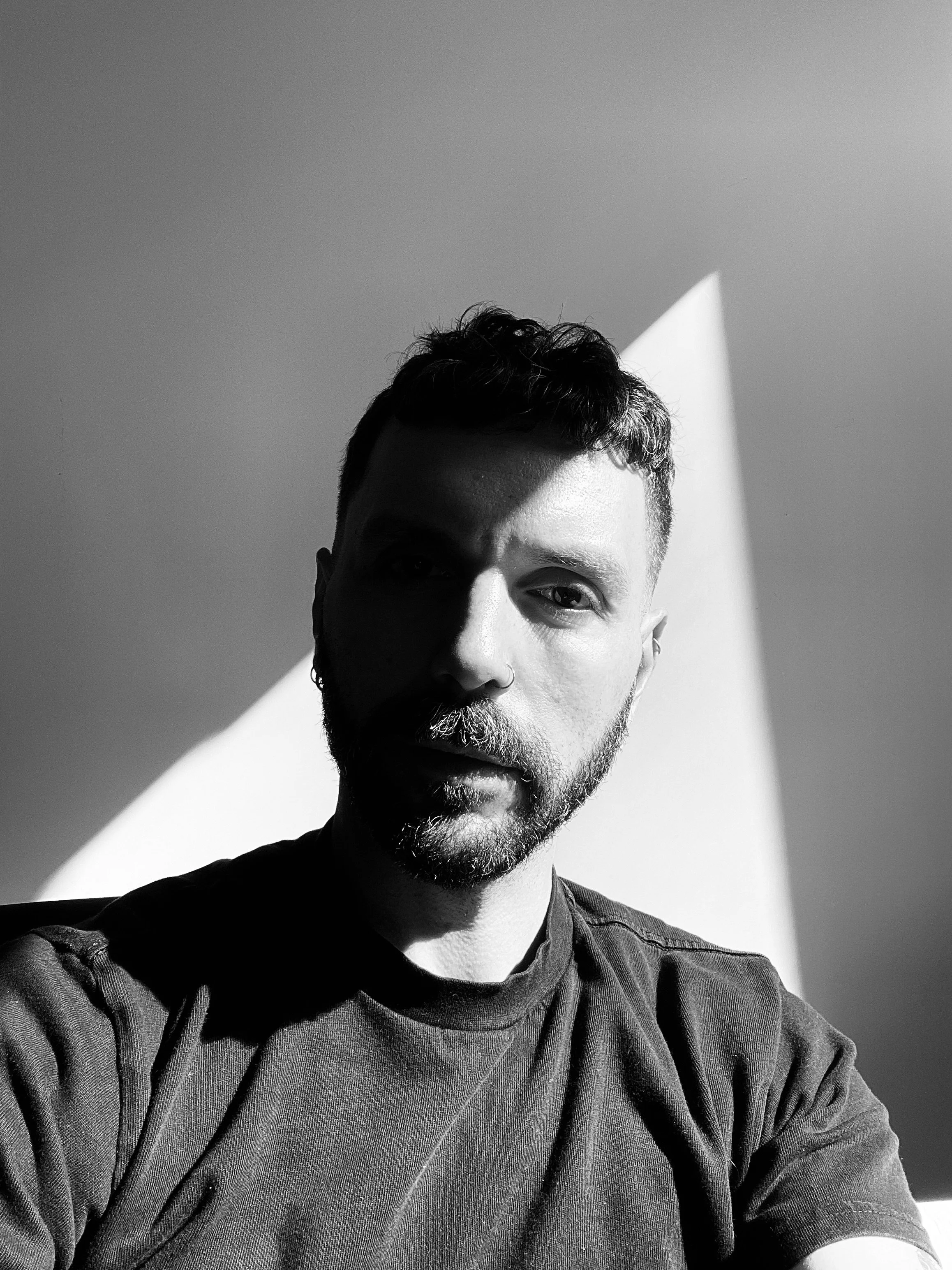Black and white photo of a man with short hair and a beard, wearing a t-shirt, sitting near a window with sunlight creating shadows on his face.
