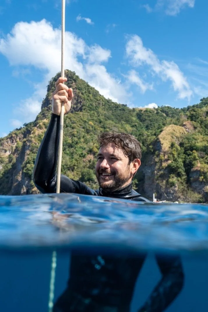 A man in a wetsuit holding a rope while floating in the water near a lush green mountainous coastline under a partly cloudy sky.