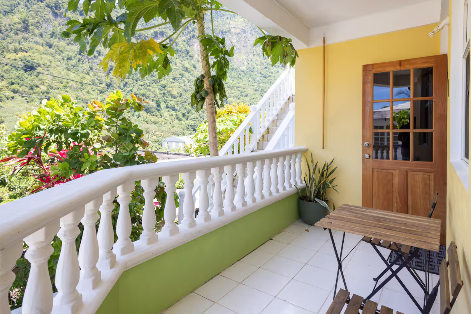 Balcony with a white railing, yellow wall, wooden door with glass panels, green plant in a pot, and outdoor furniture, overlooking a lush green mountain landscape.