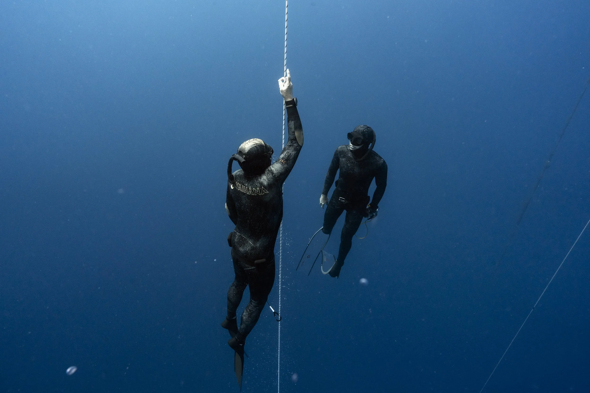 Two scuba divers underwater holding a jump rope, with one person directly holding one end and the other person holding the other end, both wearing wetsuits, masks, and fins.