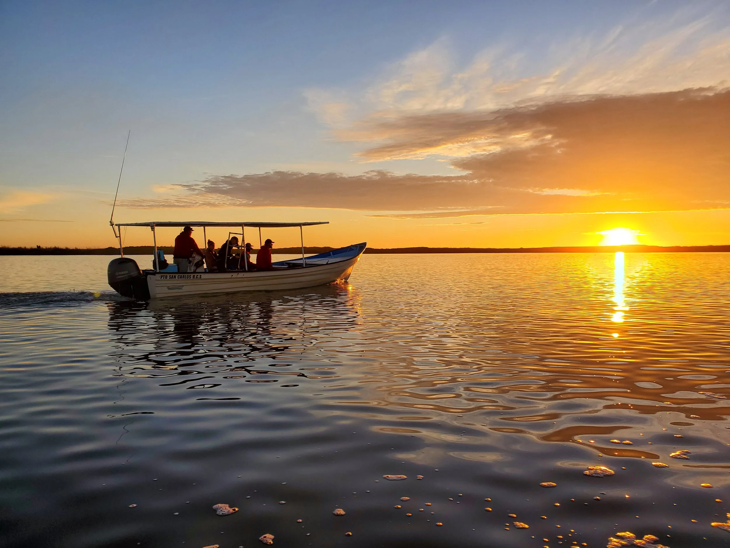 A boat with several people onboard glides across a body of water at sunset, with the sky painted in shades of orange, yellow, and blue, and the sun nearing the horizon.
