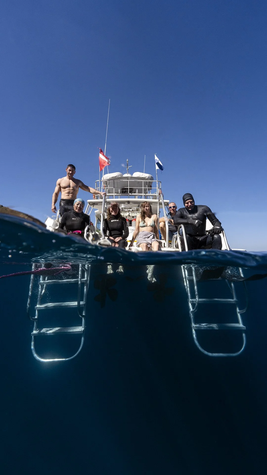 Group of six people on a boat, some in wetsuits, with a clear blue sky and ocean surrounding them.