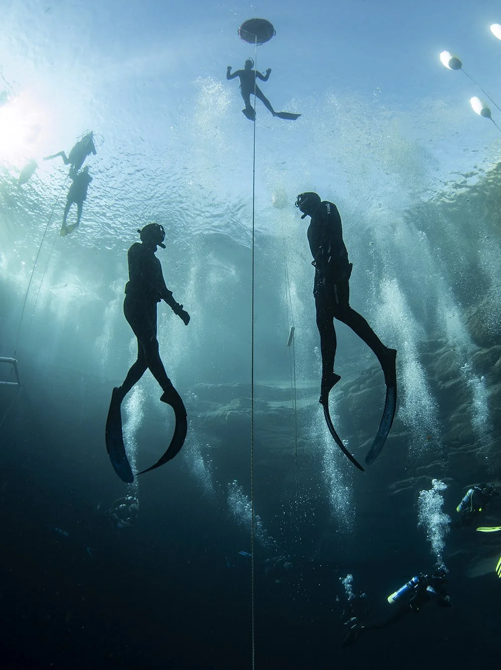 Underwater scene with two scuba divers and two freedivers, one freediver holds onto a rope with fins, surrounded by bubbles in a large pool or ocean environment.