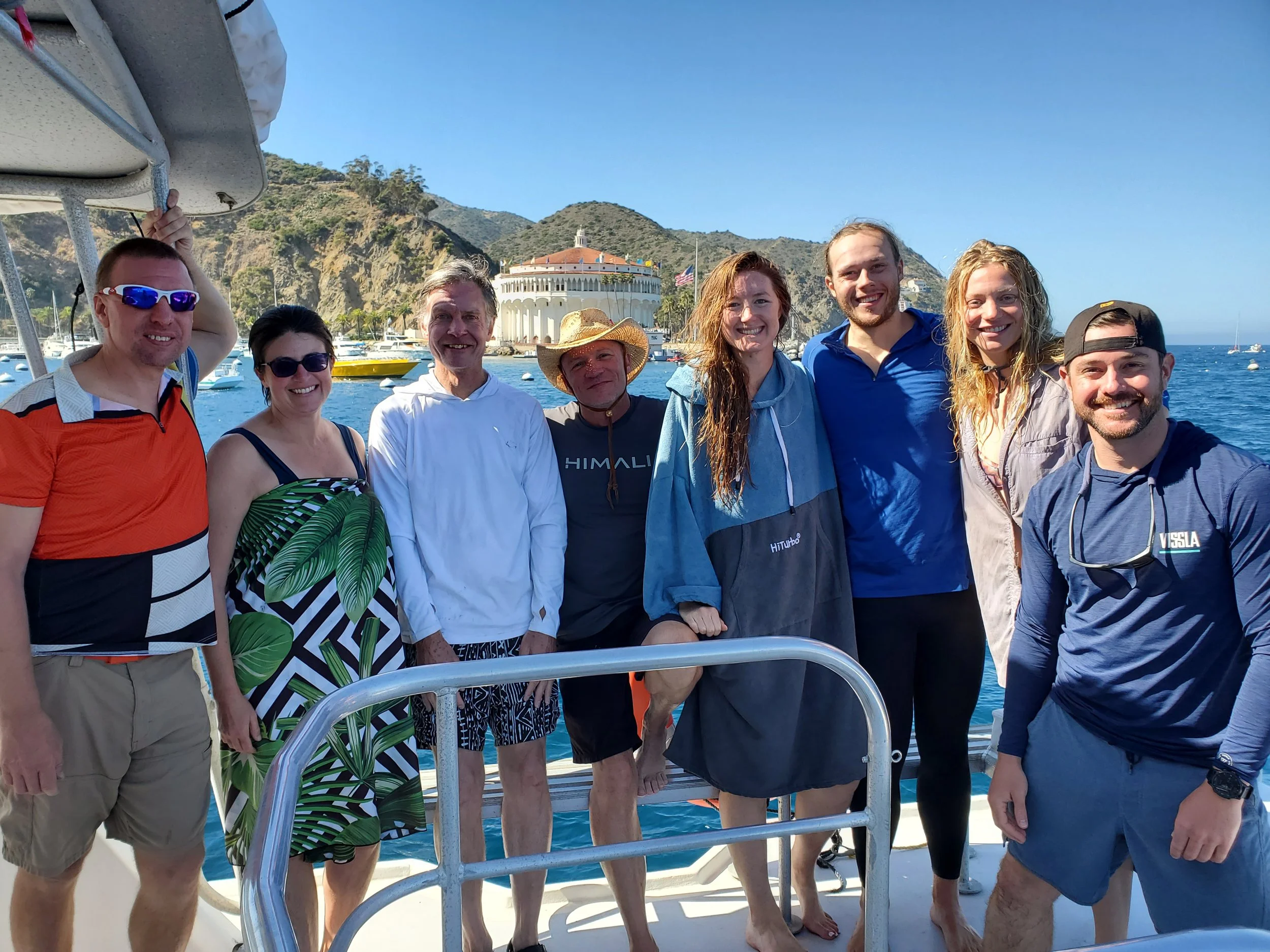 Group of eight people posing on a boat with a scenic harbor, mountains, and a building with a red-tiled roof in the background, sunny day with clear sky.