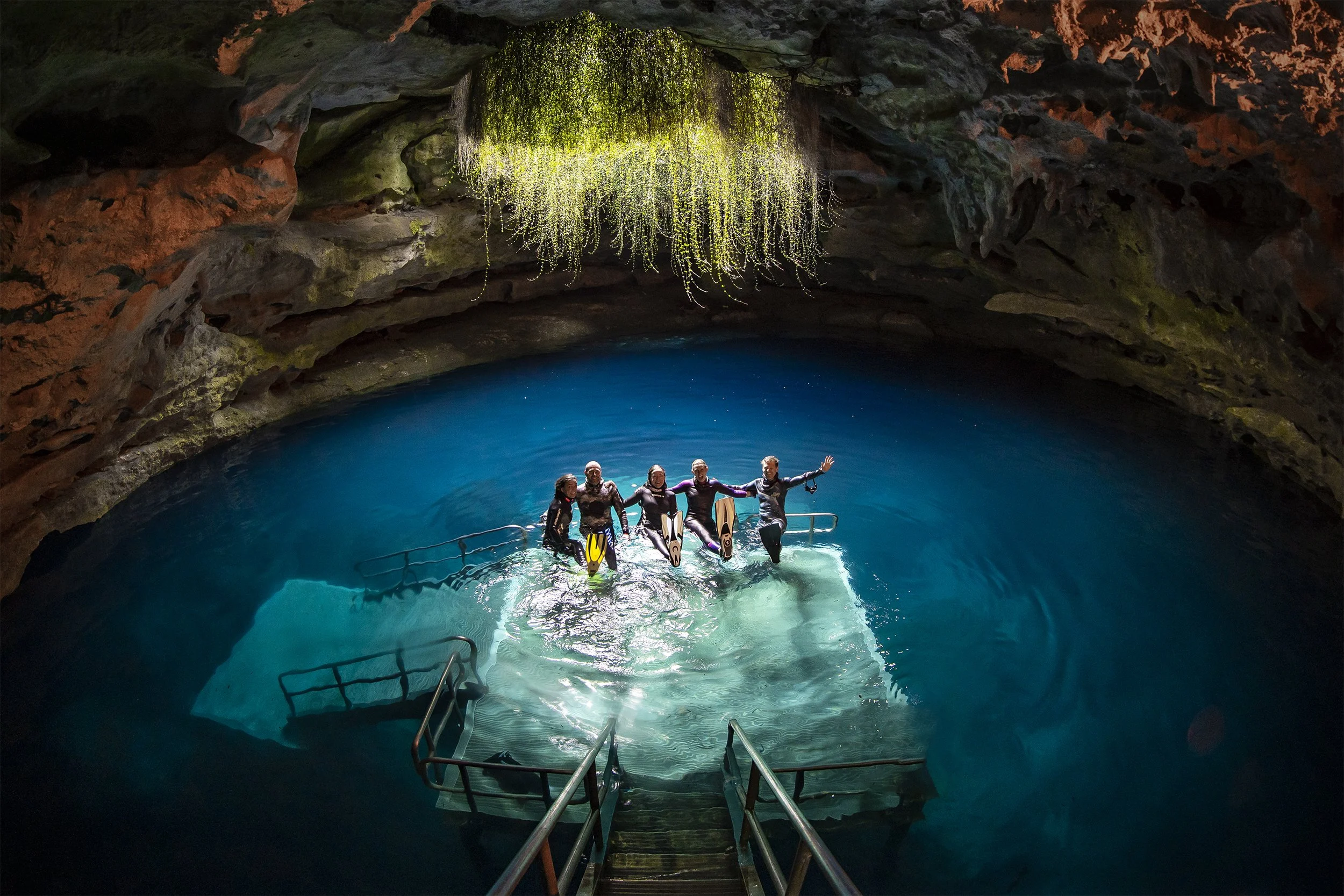 Group of five people holding hands and standing in a swimming pool inside a cave, with rocky walls and a large, glowing hanging moss or plant overhead.