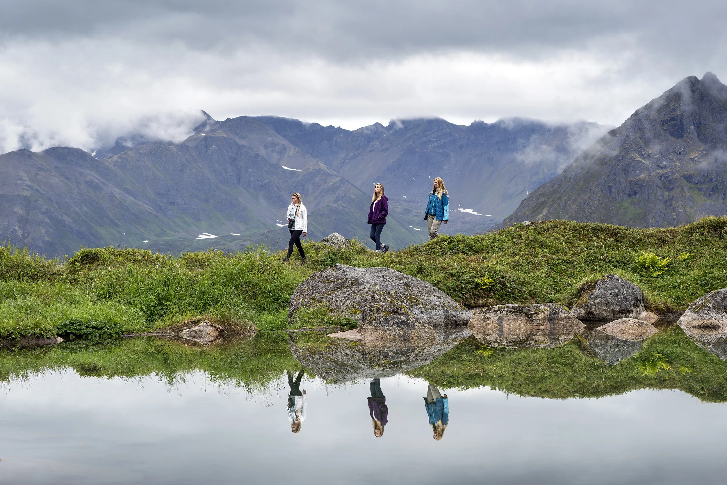 Three women hiking in a mountainous landscape with overcast skies, standing on green grass with rocks, reflected in a calm body of water in the foreground.