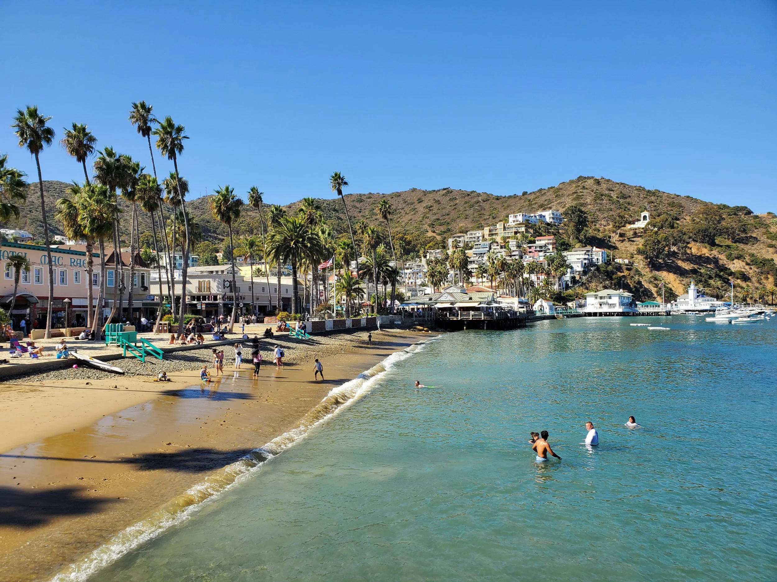 People swimming and relaxing on a sandy beach with palm trees, buildings, and hillside in the background under a clear blue sky.