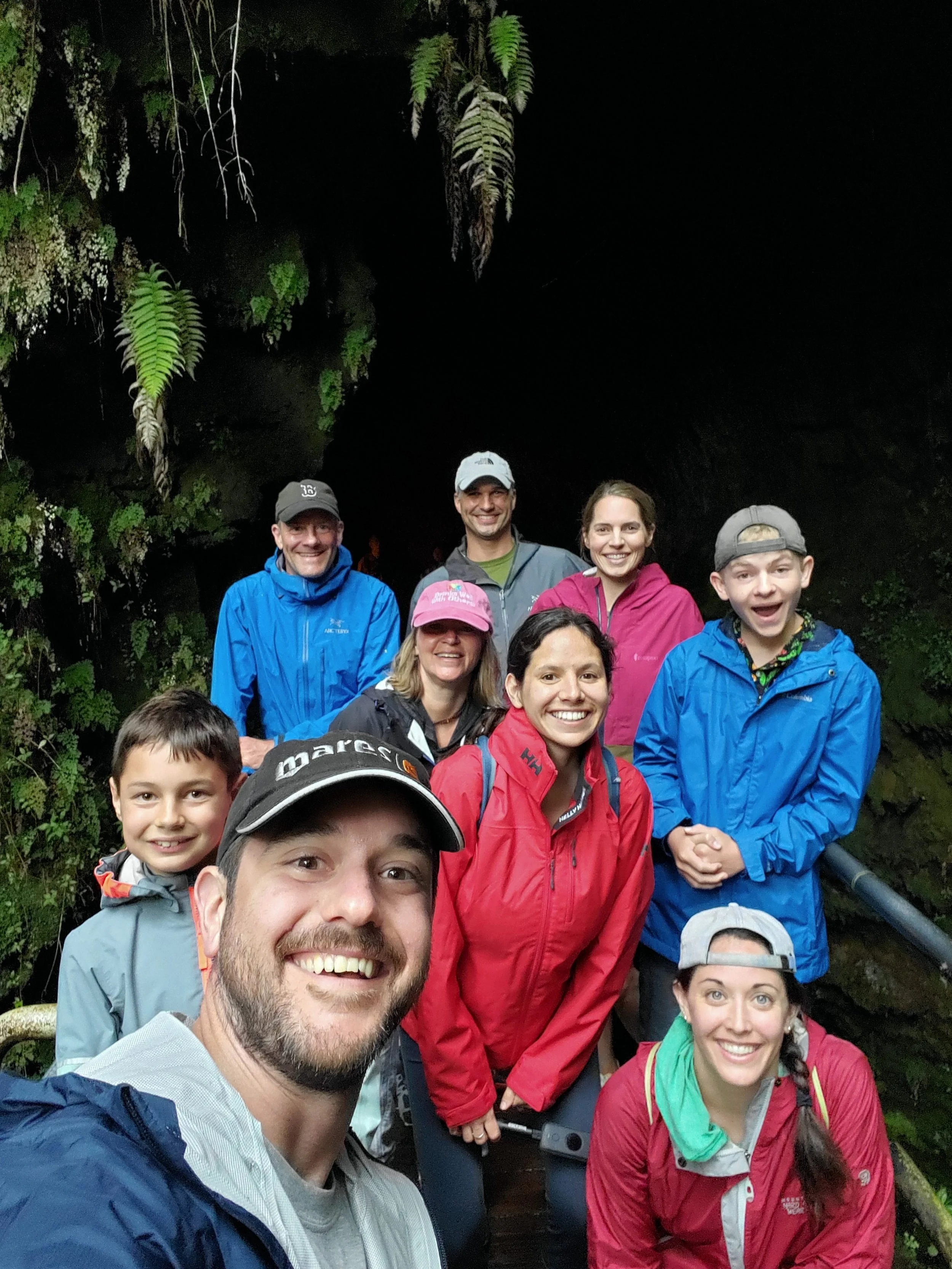 A group of people smiling and posing for a photo in a dark, lush, outdoor setting with hanging ferns and foliage.