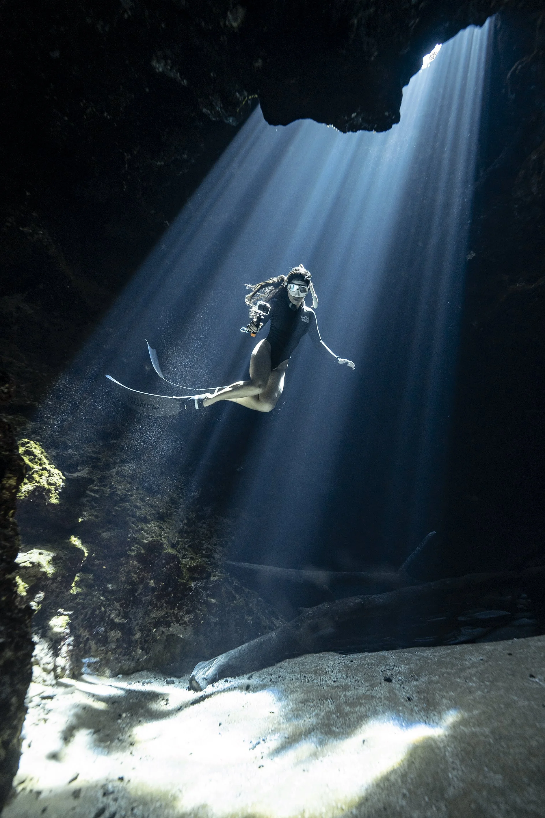 A woman in snorkeling gear swimming in a dark underwater cave illuminated by beams of sunlight from above.