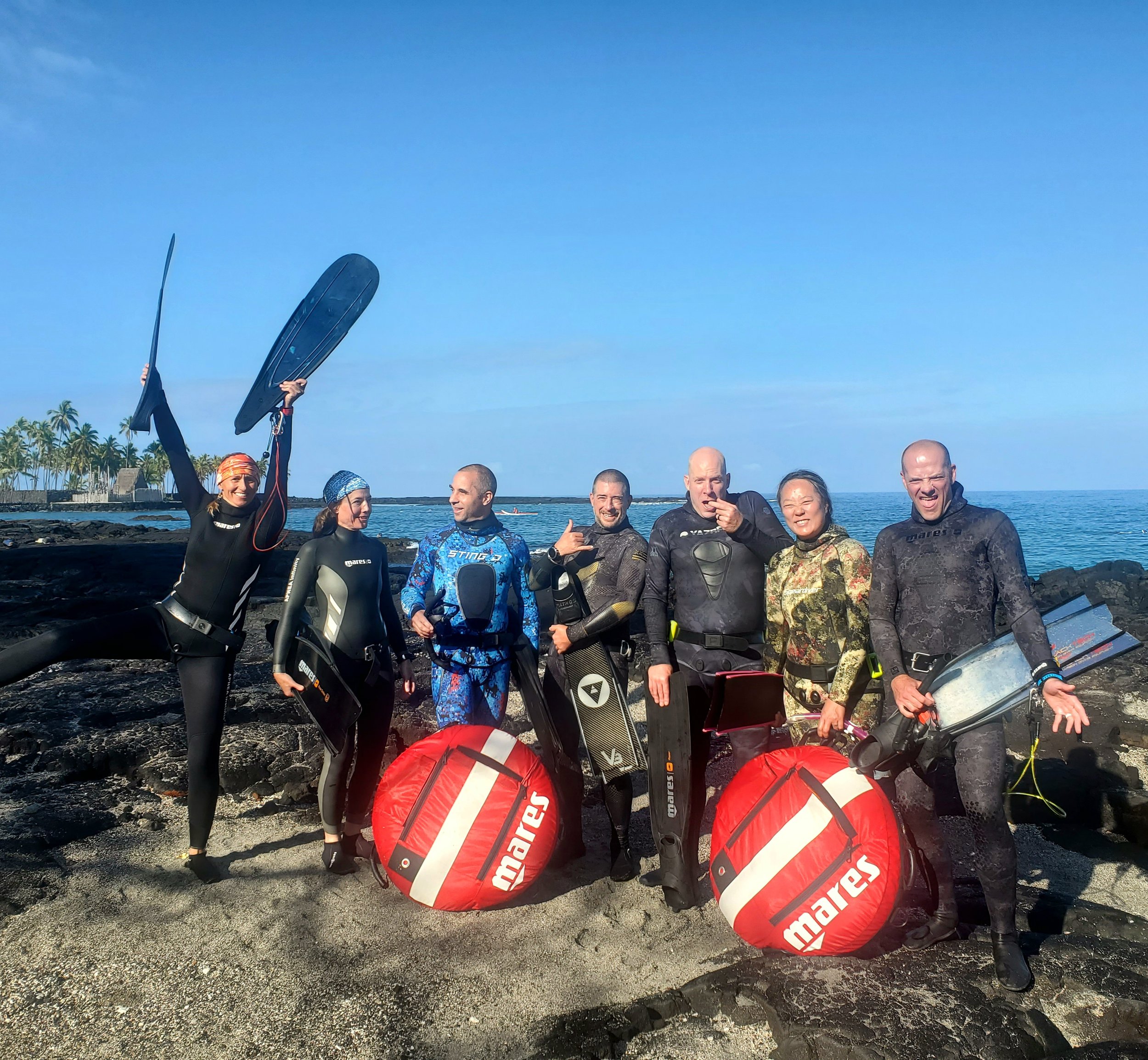 Group of people standing on rocky beach holding wakeboarding equipment, with water and palm trees in the background under a clear blue sky.