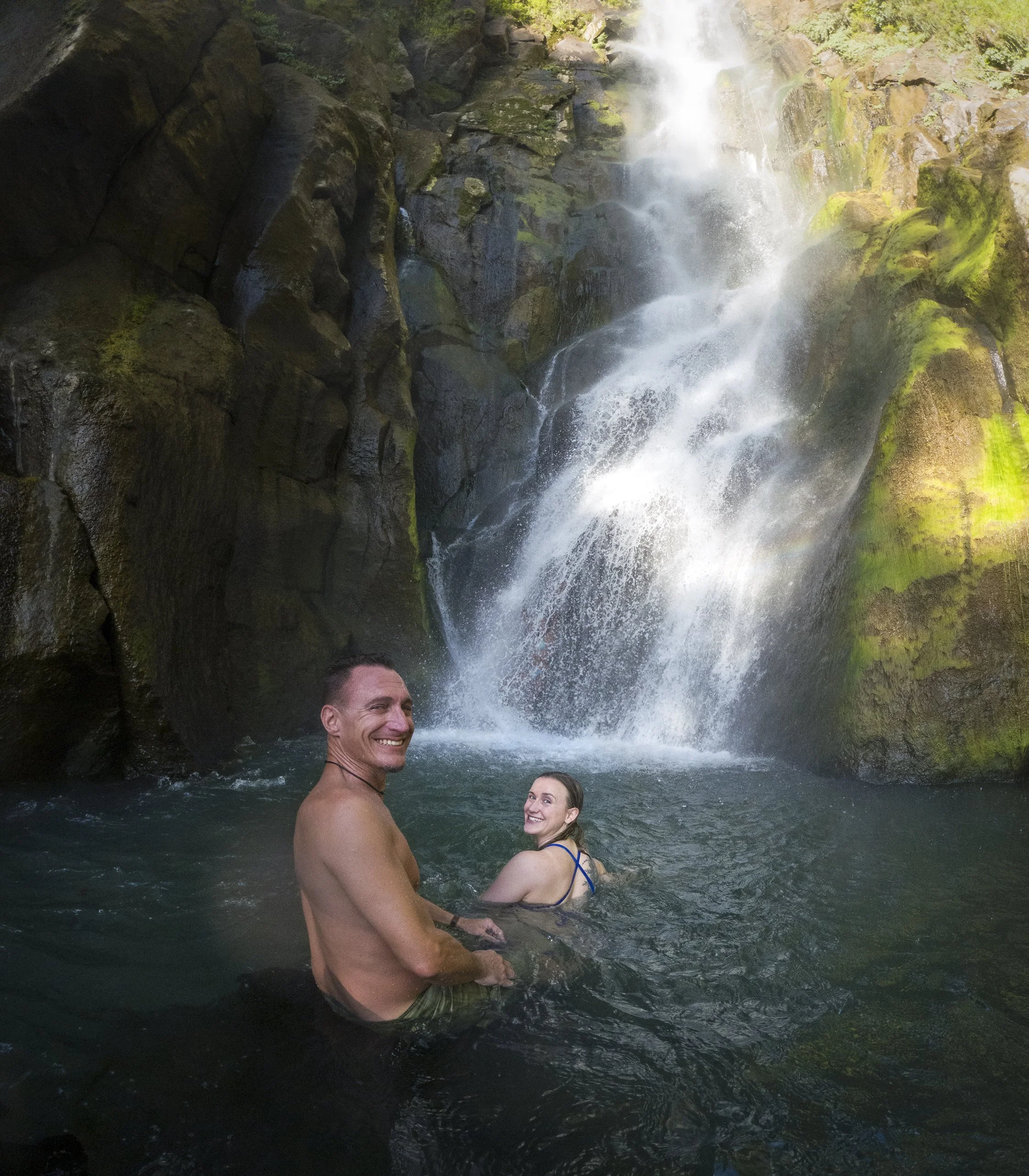 Two people, a man and a woman, are sitting in a natural water pool at the base of a waterfall in a lush green forest. The man is shirtless and has a smile, while the woman is wearing a swimsuit and is smiling back.