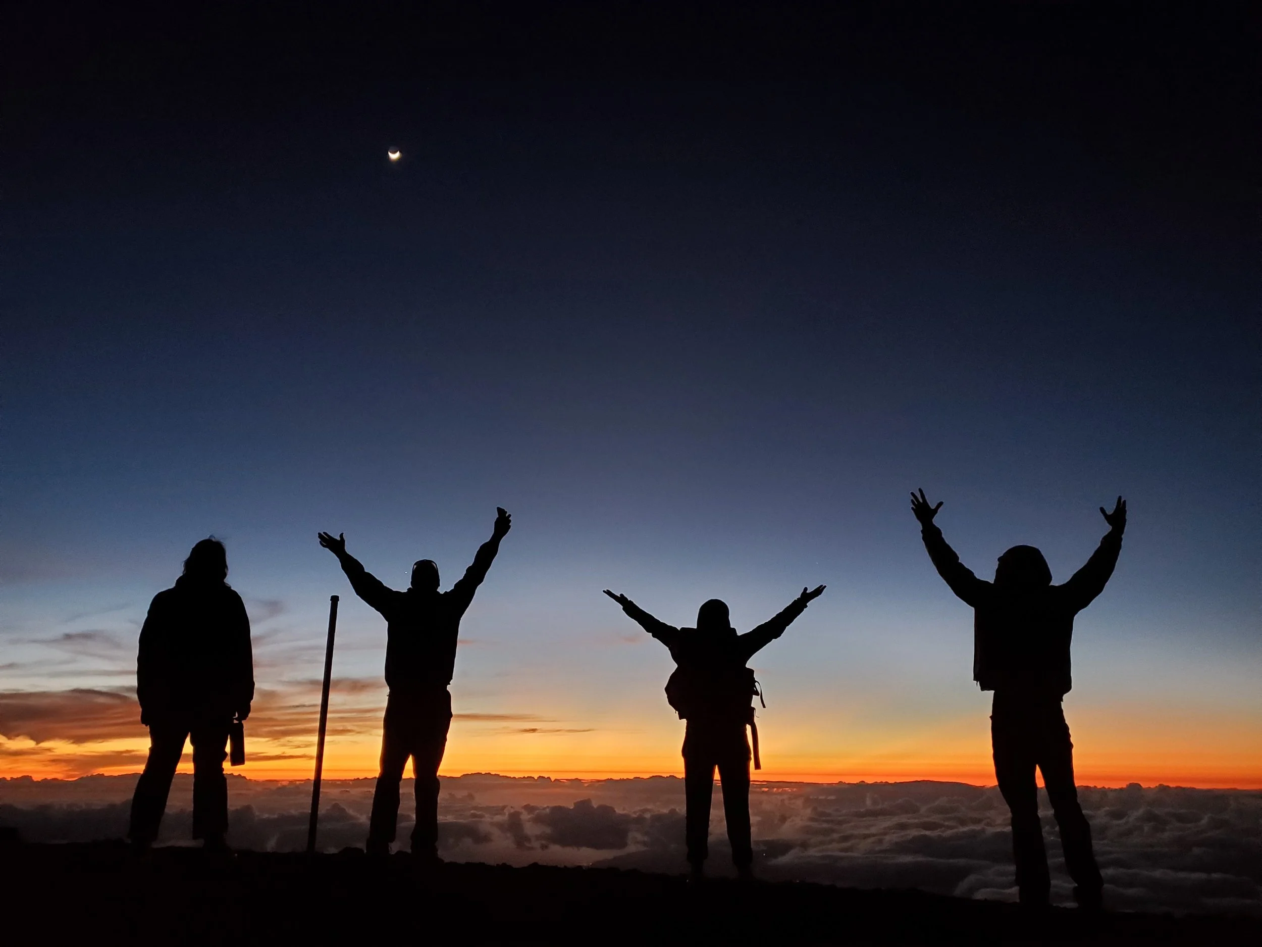 Silhouettes of six people standing outdoors during sunset, with some raising their hands, against a sky with a small crescent moon and clouds near the horizon.
