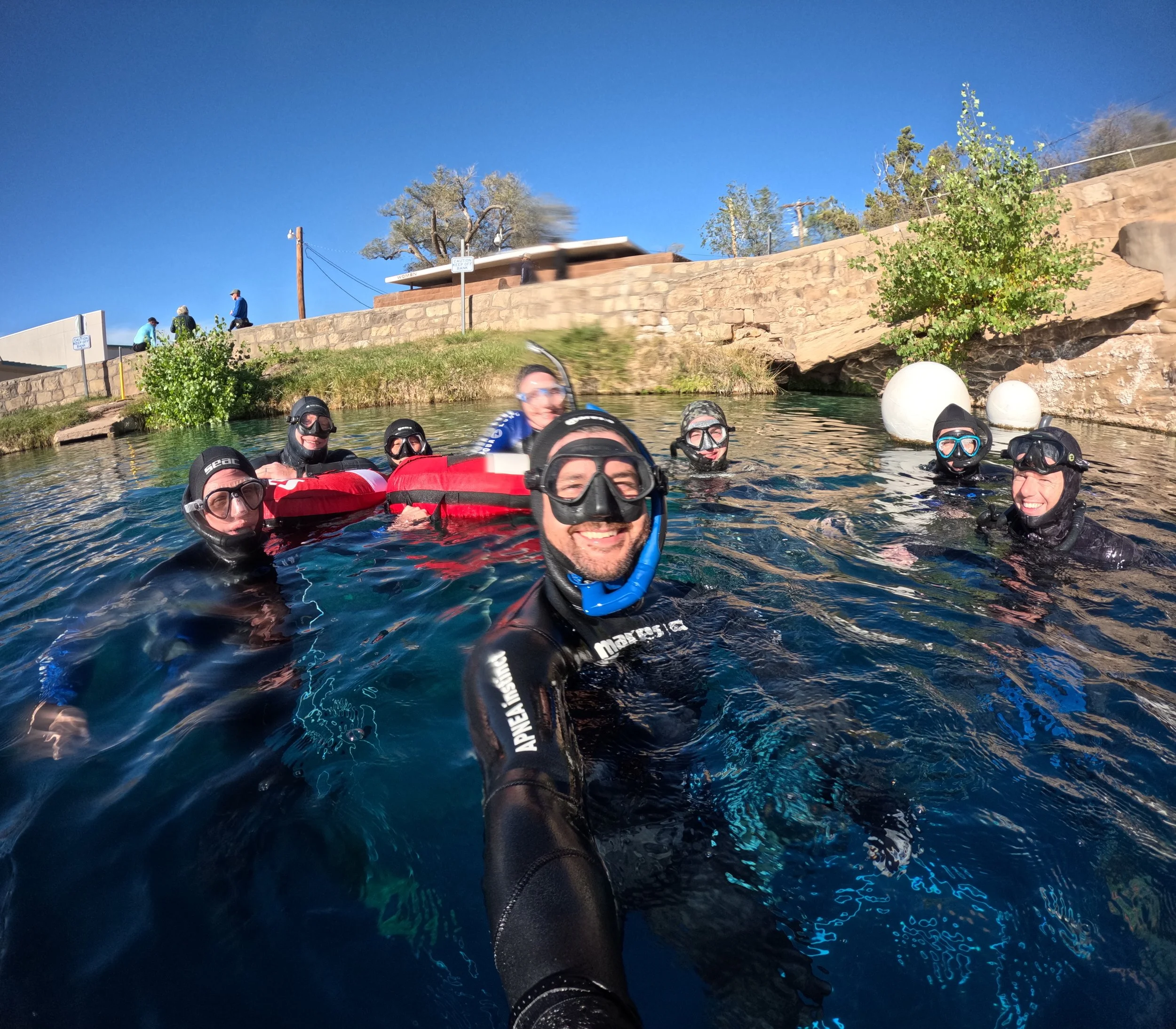 A group of people wearing wetsuits and helmets smiling and taking a selfie while swimming in a river with rocky banks and trees in the background on a sunny day.