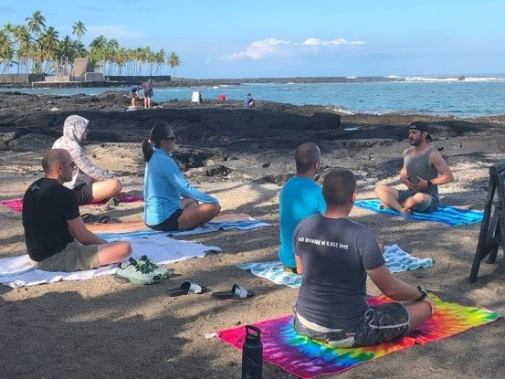 Group of people practicing meditation on the beach during the daytime with palm trees and ocean in the background.