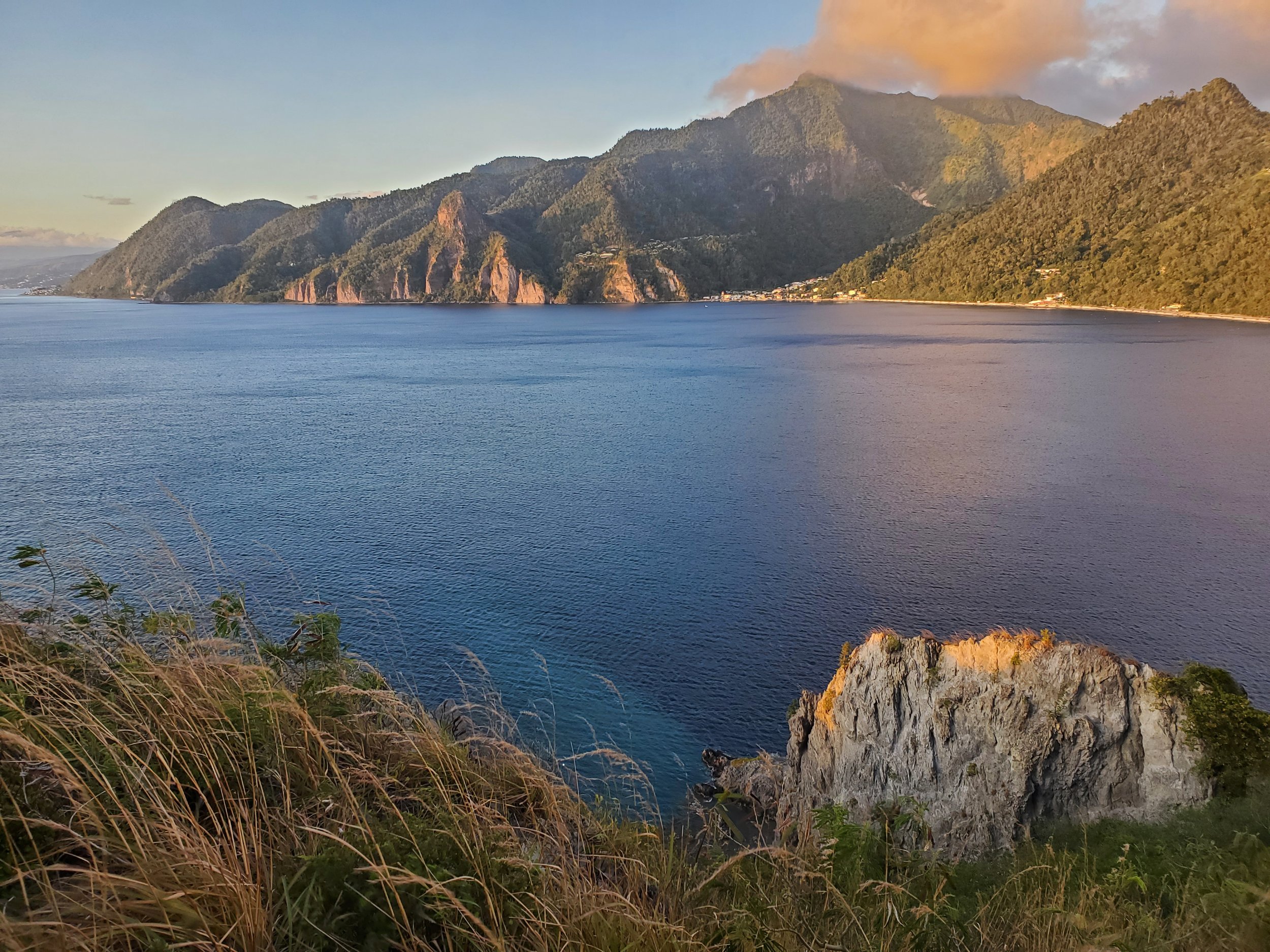 Scenic view of a large body of water with mountains in the background and a hillside covered with grasses and rocks in the foreground at sunset or sunrise.