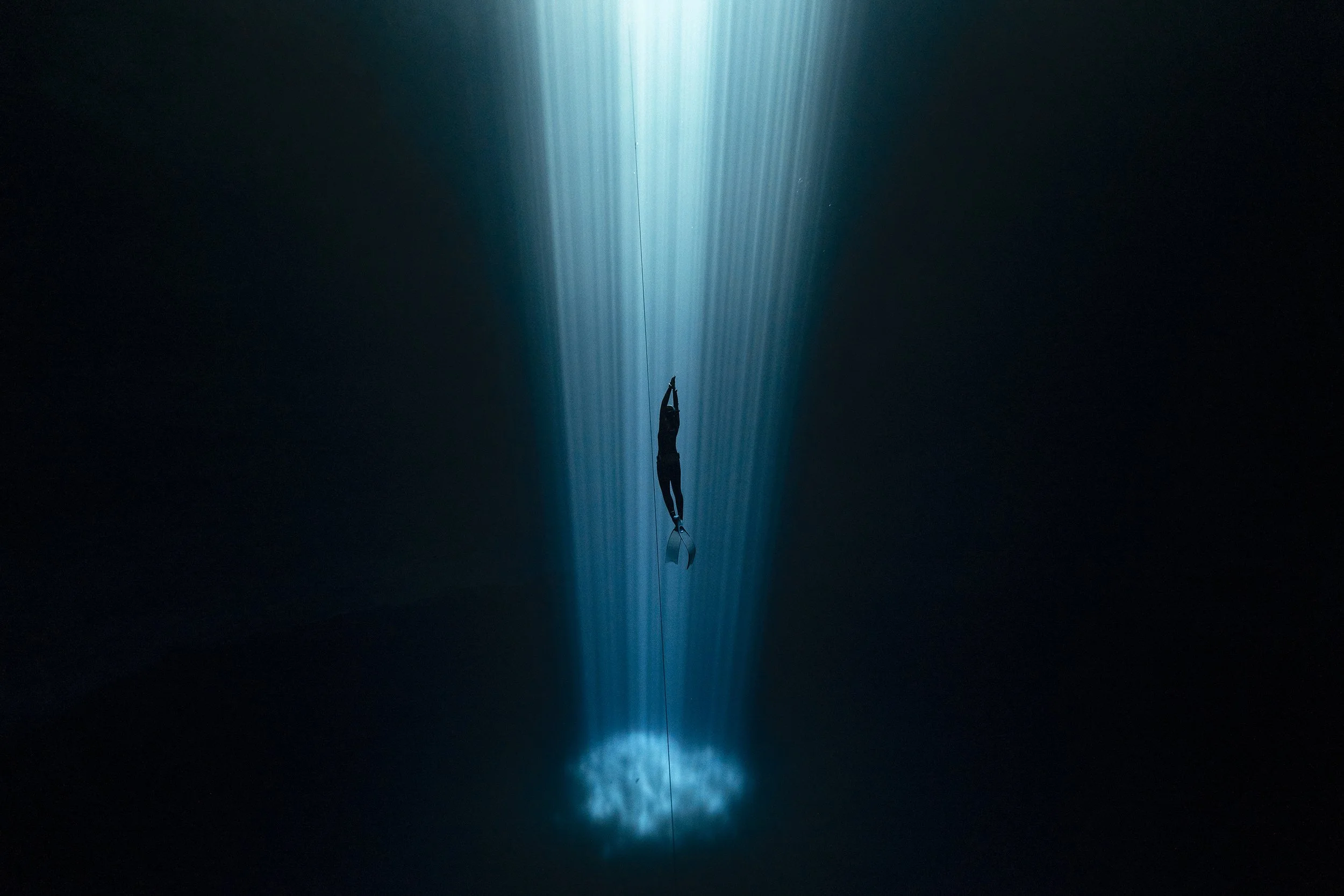 A person doing an underwater acrobatic move, with their body arched backward and arms extended, suspended above the water with blue and white light beams below.