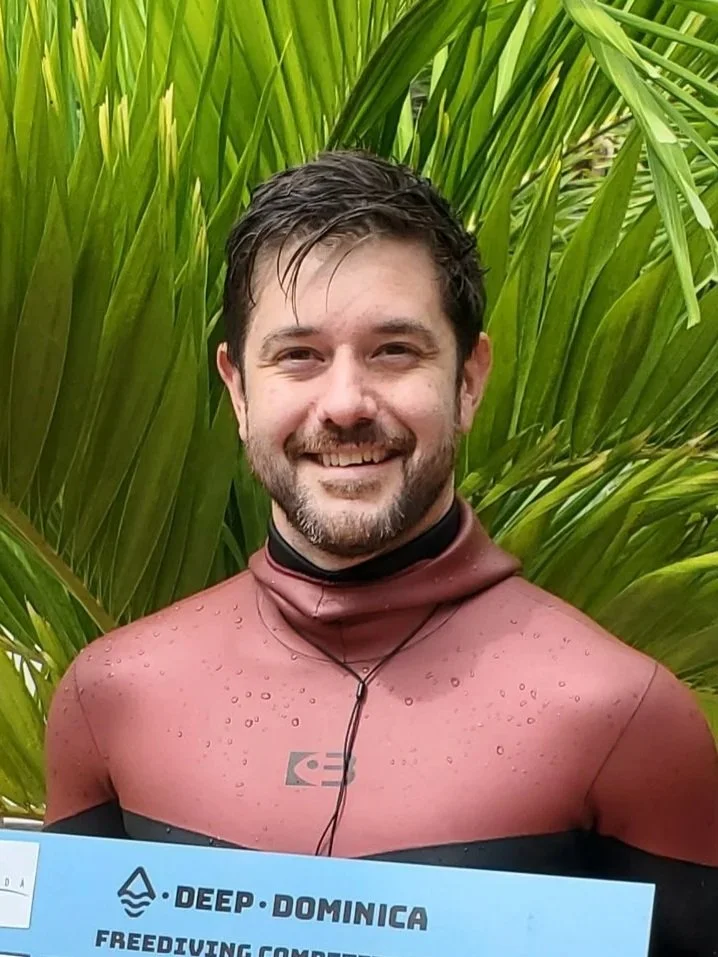 A smiling man, Drew Herrick, with dark hair and a beard, wearing a red wetsuit, standing in front of green tropical plants, holding a sign that says 'Deep Dominica Freediving Competition'.