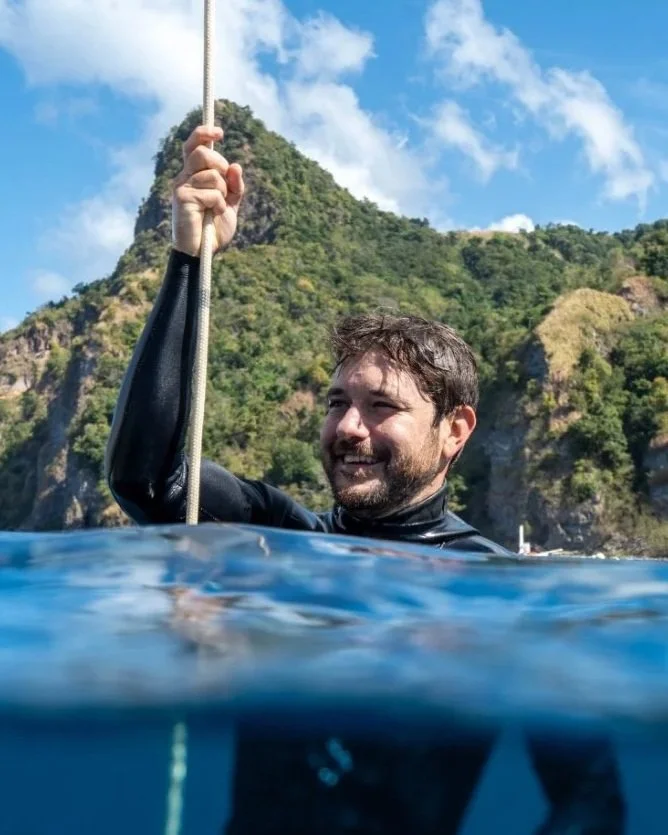 Man in a wetsuit holding onto a rope in the water near a mountainous island with a partly cloudy sky.