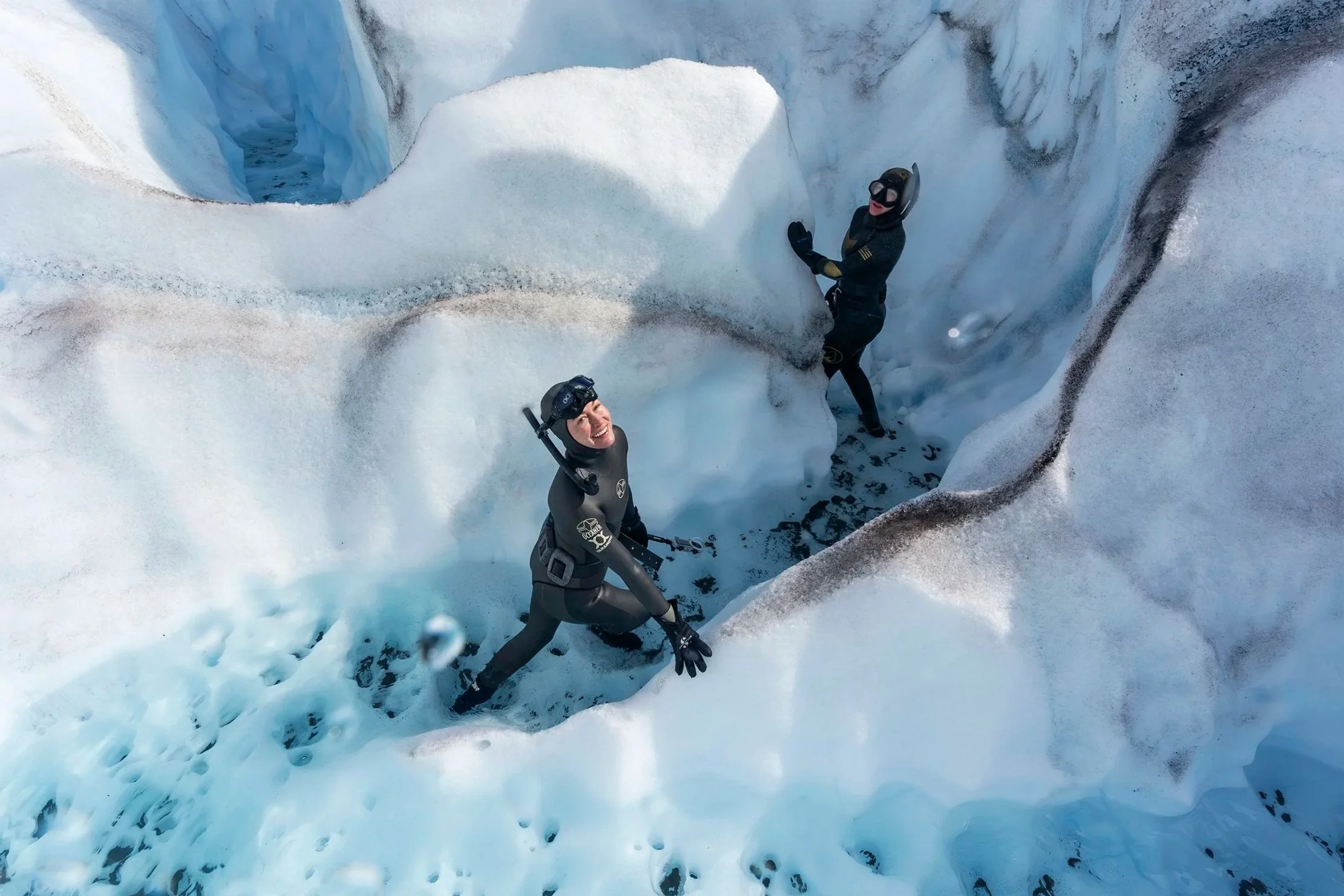 Two people in wetsuits and helmets exploring a glacier cave with icy walls and a deep blue ice opening.