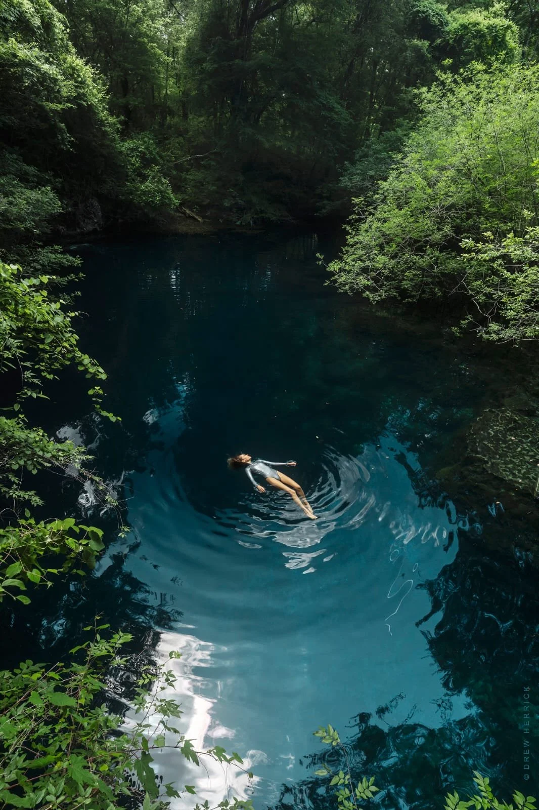 A person floating peacefully in a deep blue river surrounded by lush green trees.
