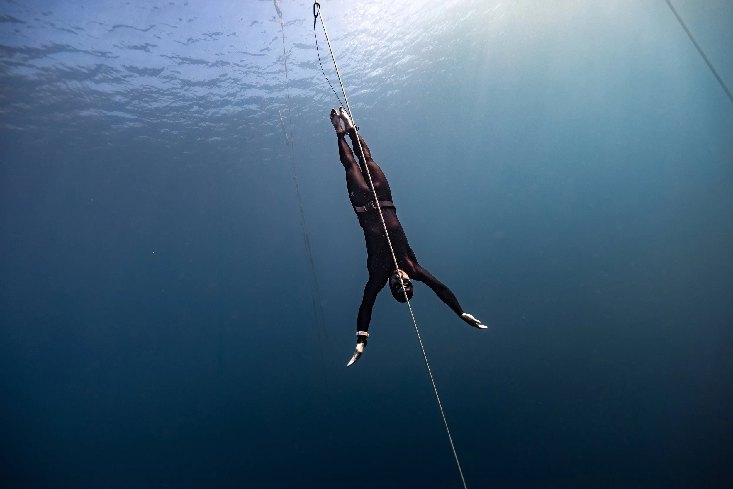 A person performing a trapeze stunt underwater, hanging upside down from a trapeze bar with legs spread apart, wearing black wetsuit, with water and light visible in the background.