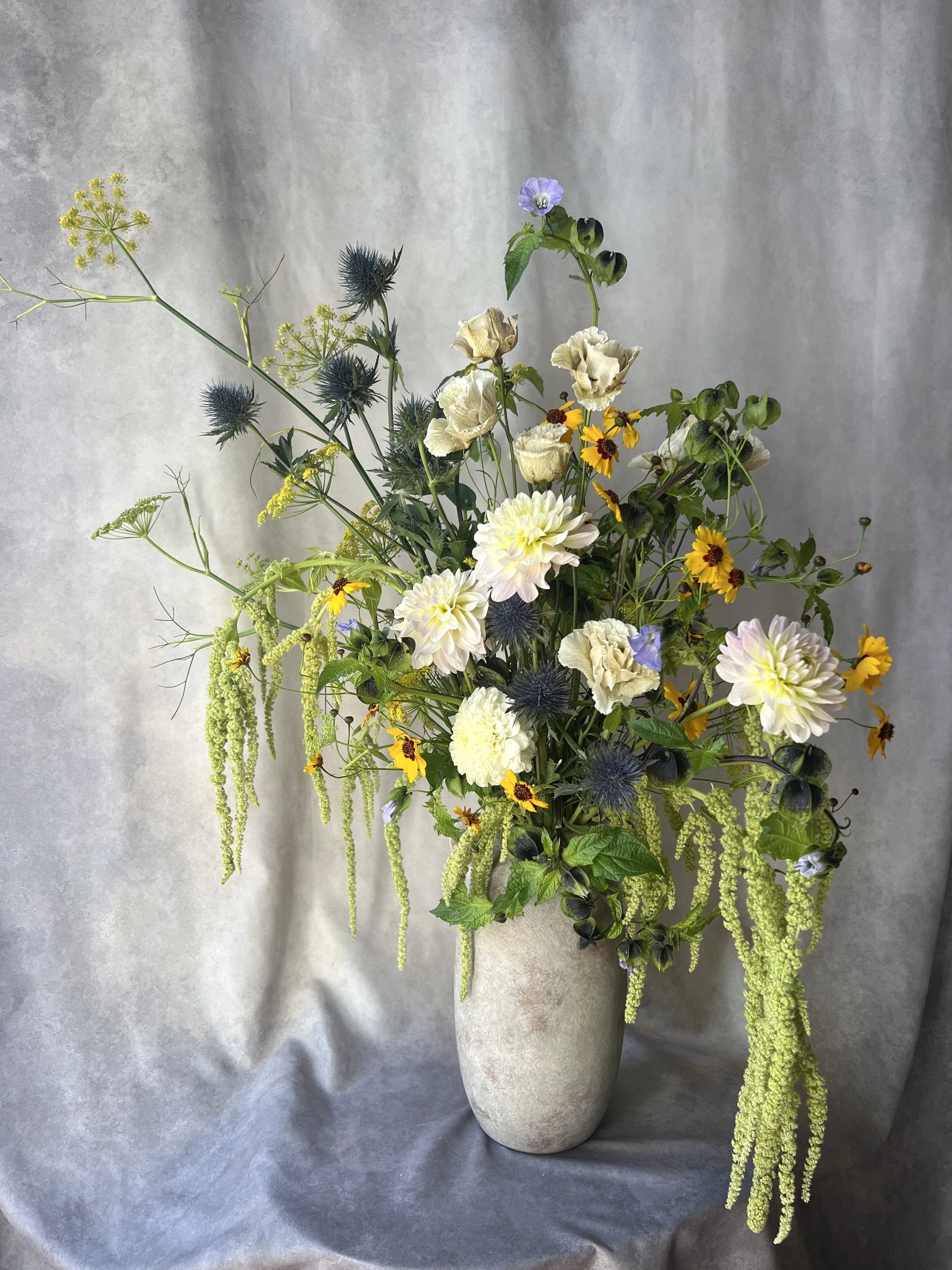 Loose, garden-inspired floral arrangement with white, yellow and blue seasonal blooms in a tall ceramic vase.