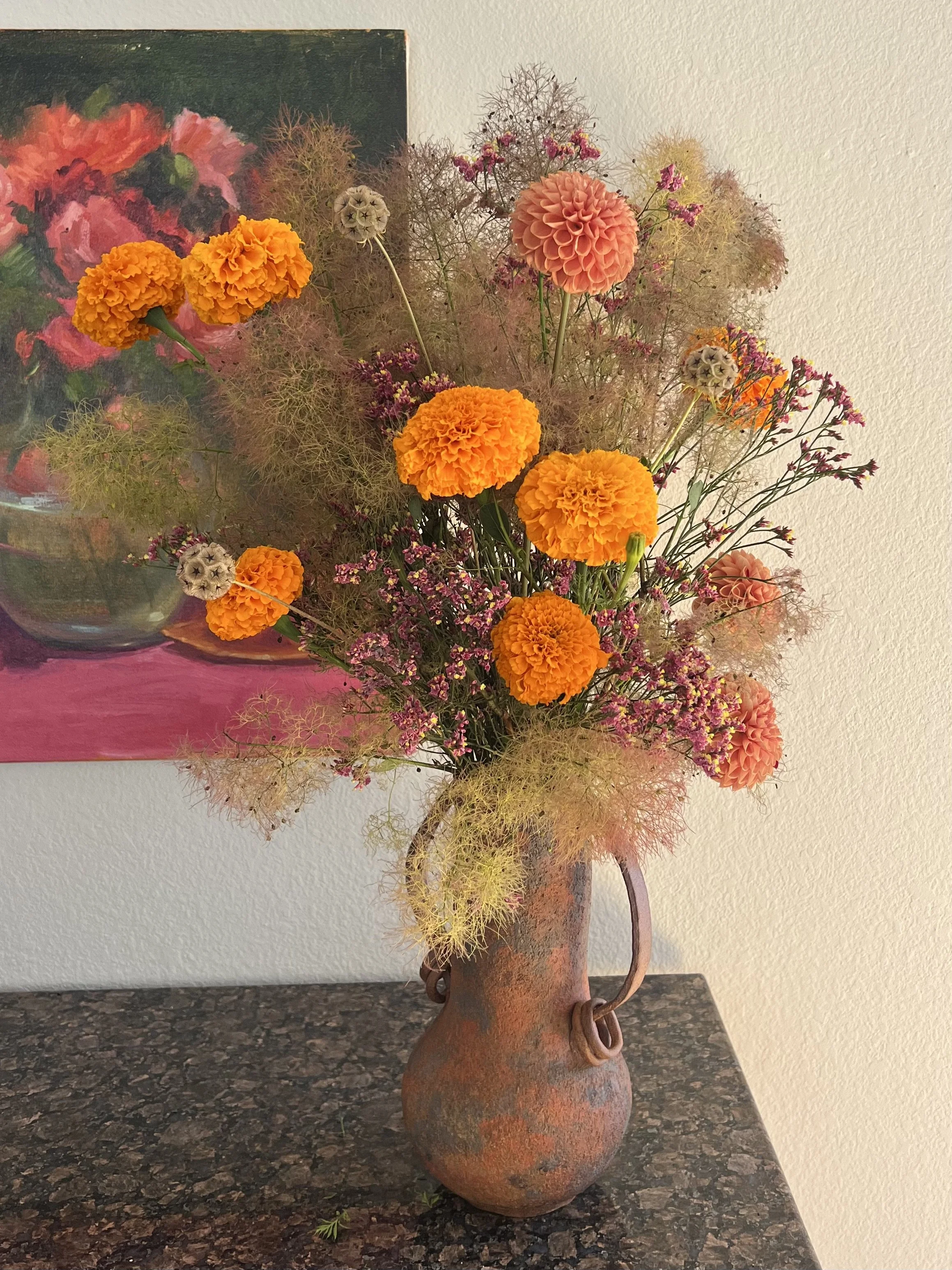 Seasonal floral arrangement with orange marigolds, dahlias and airy pink blooms in a handmade clay vase.