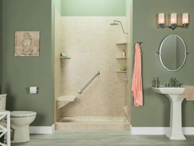A bathroom with a walk-in shower, a toilet, a small white cabinet, and a pedestal sink. The walls are painted green, with a framed artwork on the left and three vanity lights above the mirror on the right. The shower has beige tiles, a handheld showerhead, shelves with toiletries, and a pink towel hanging beside it.