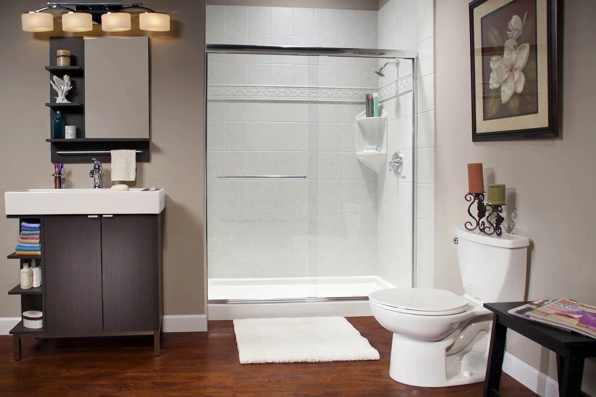 Bathroom with a walk-in glass shower, white toilet with decorative candles on top, dark wood vanity with towels and toiletries, framed artwork on the wall, and a wooden floor.