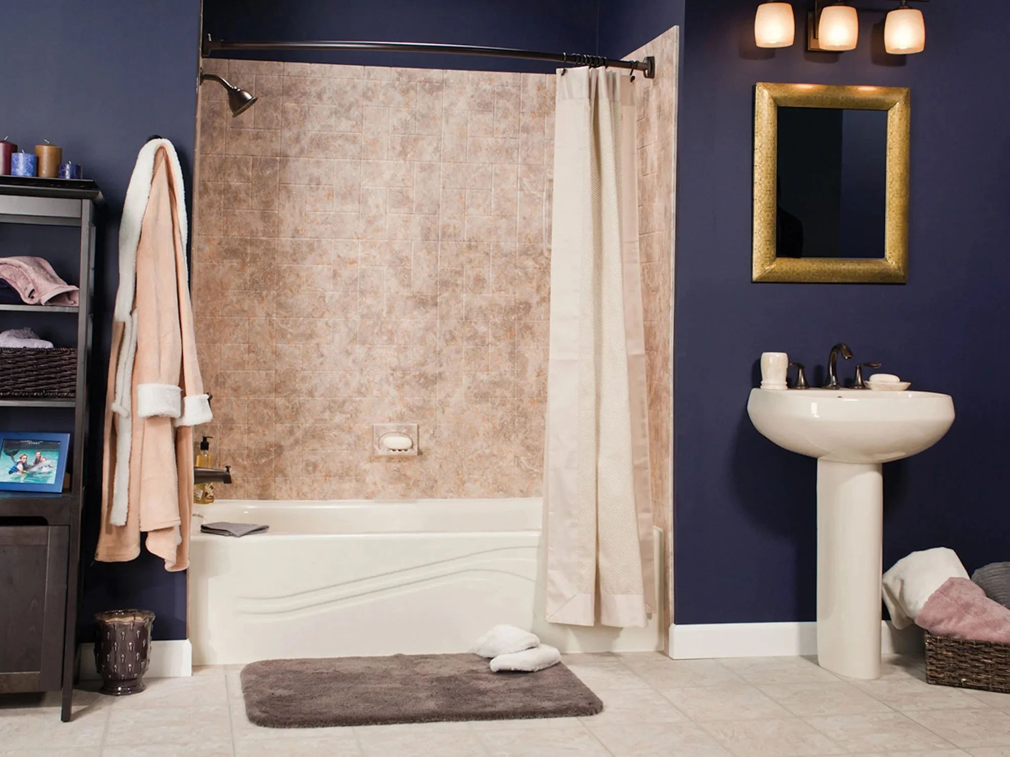 Bathroom with beige tile bathtub, navy blue wall, white pedestal sink, framed mirror, light fixture, shelving with towels and toiletries, bathrobe hanging, gray bath mat, and laundry basket.