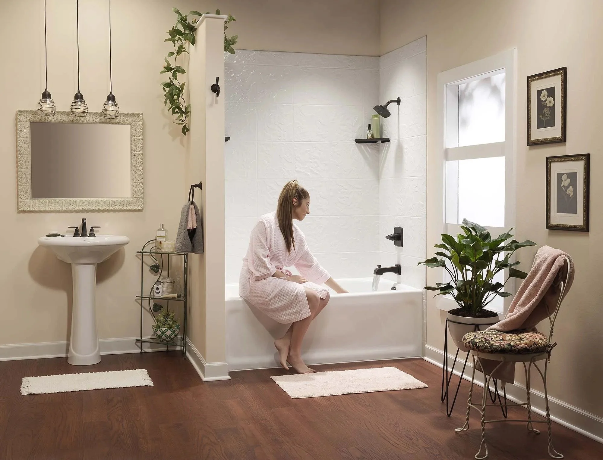 A woman in a pink bathrobe sitting on the edge of a bathtub in a cozy bathroom. The bathroom has wooden floors, a small sink with a mirror above, a metal shelf with plants and toiletries, a window, and framed floral artwork. There is a plant and a ch
