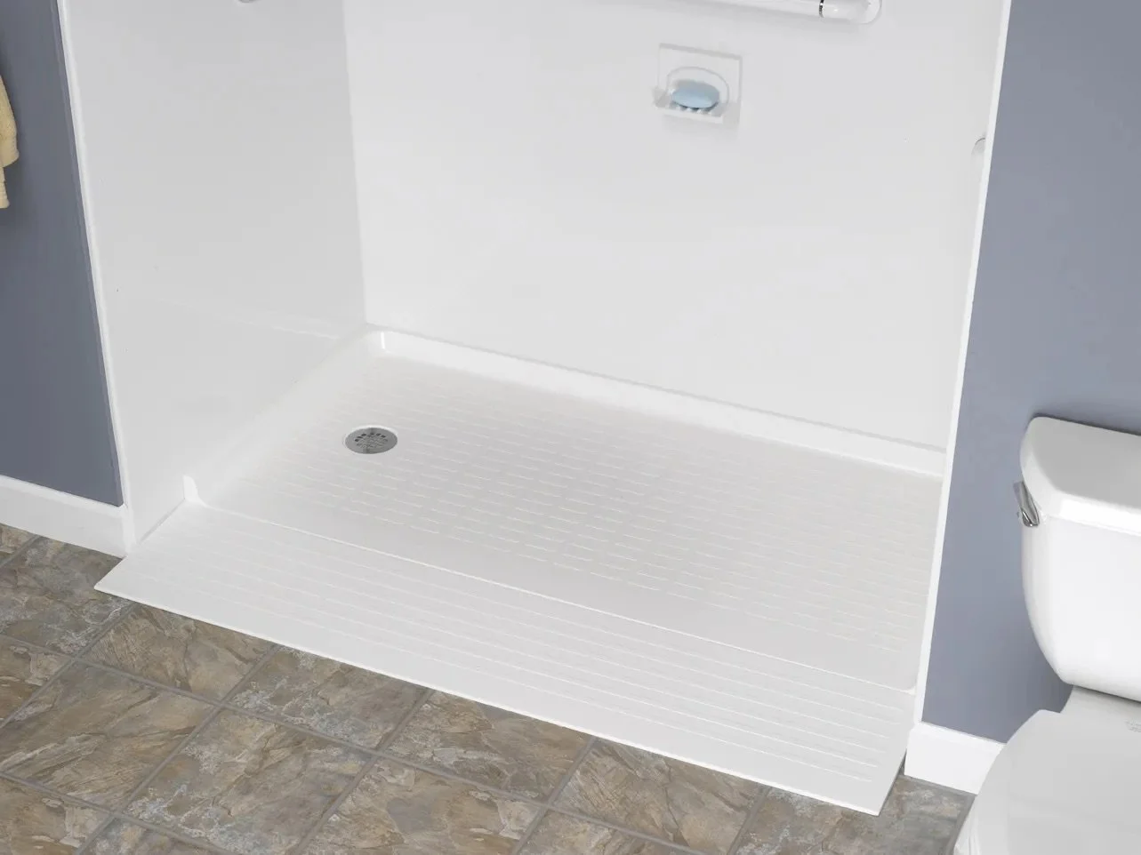 Empty white shower with drain, next to a toilet on a tiled bathroom floor, with a dark gray wall on the right.