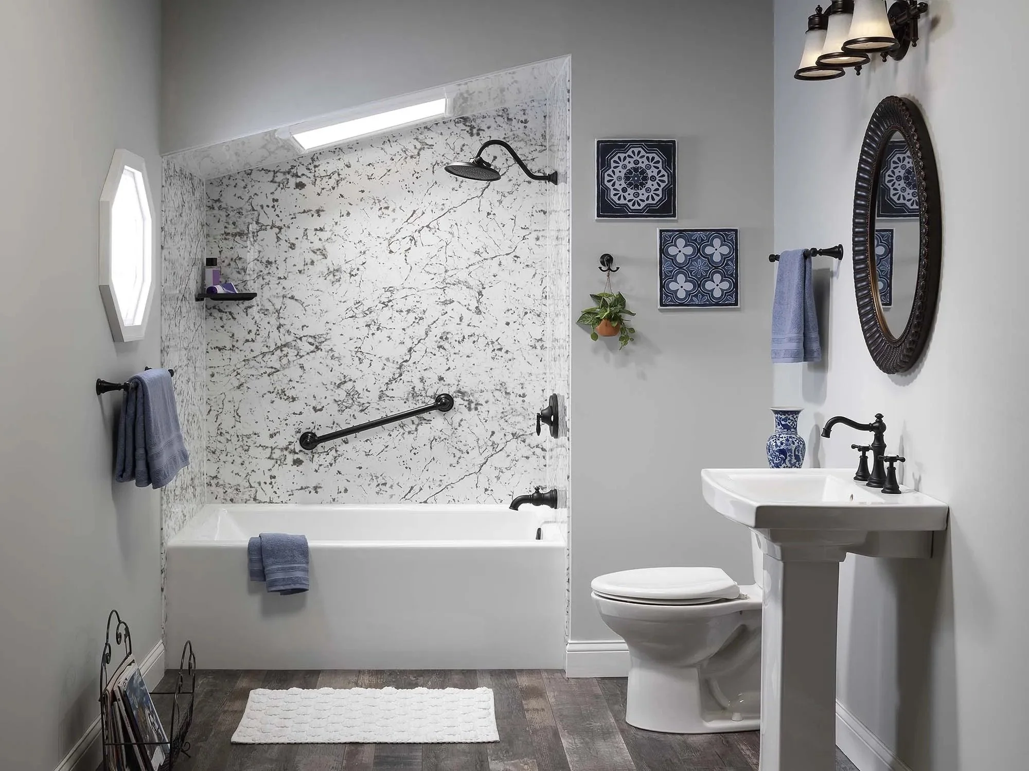 Bathroom with a white bathtub, black fixtures, a pedestal sink, mirror, toilet, and blue towels, with patterned tiles on the wall and framed artwork, a hanging plant, and a bath mat on dark wood flooring.