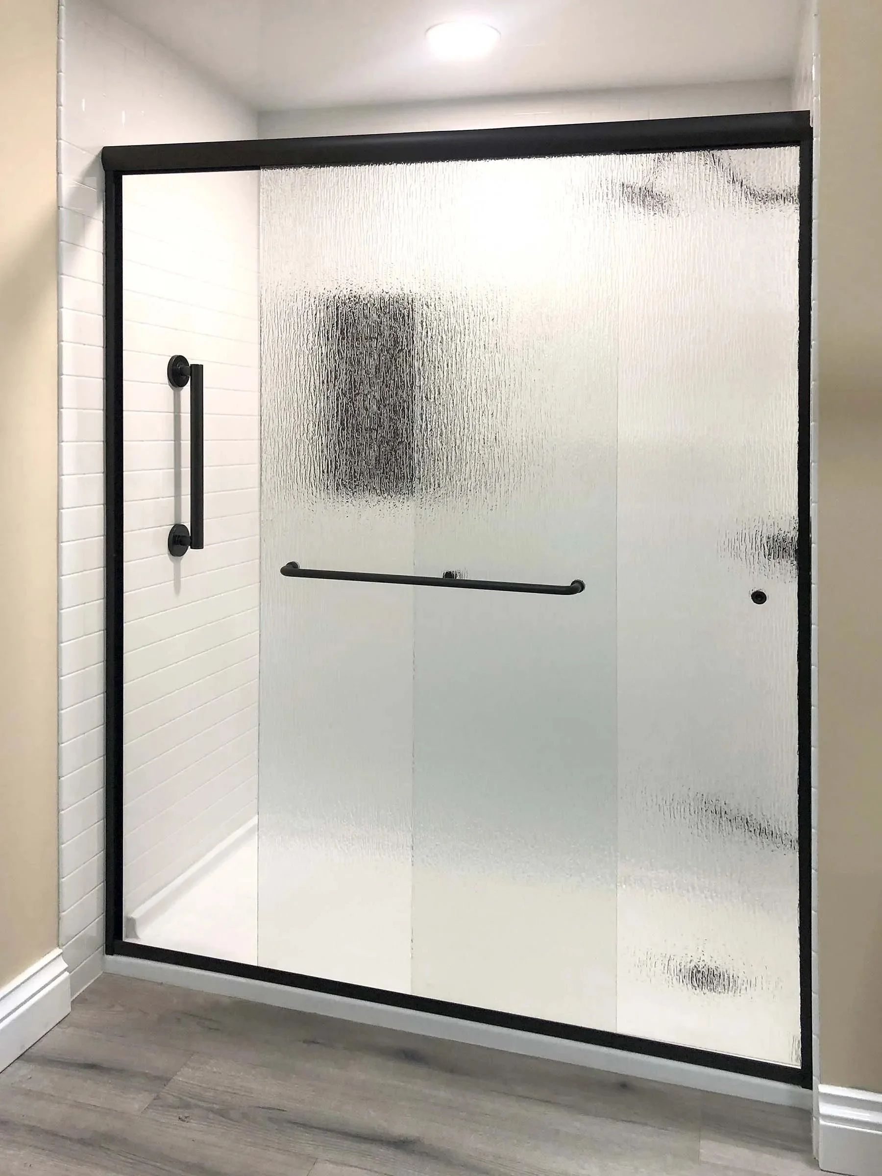 A walk-in shower with frosted glass doors, black handles, and a white shower base. The shower has white subway tile walls and is located in a modern bathroom with wood-look flooring.